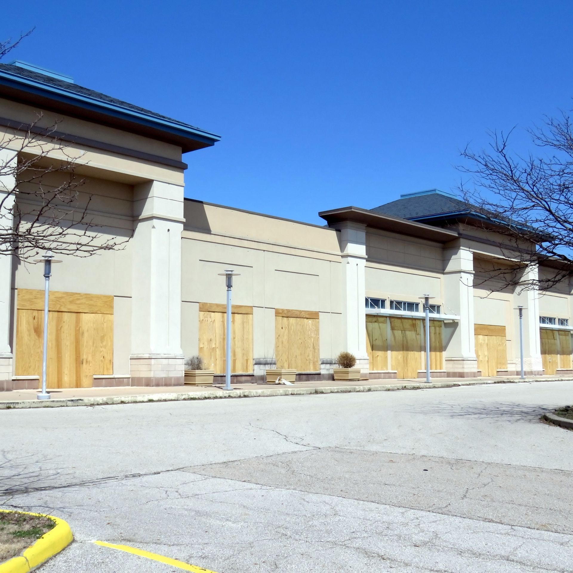 Boarded-up storefronts in a strip mall parking lot under a clear blue sky