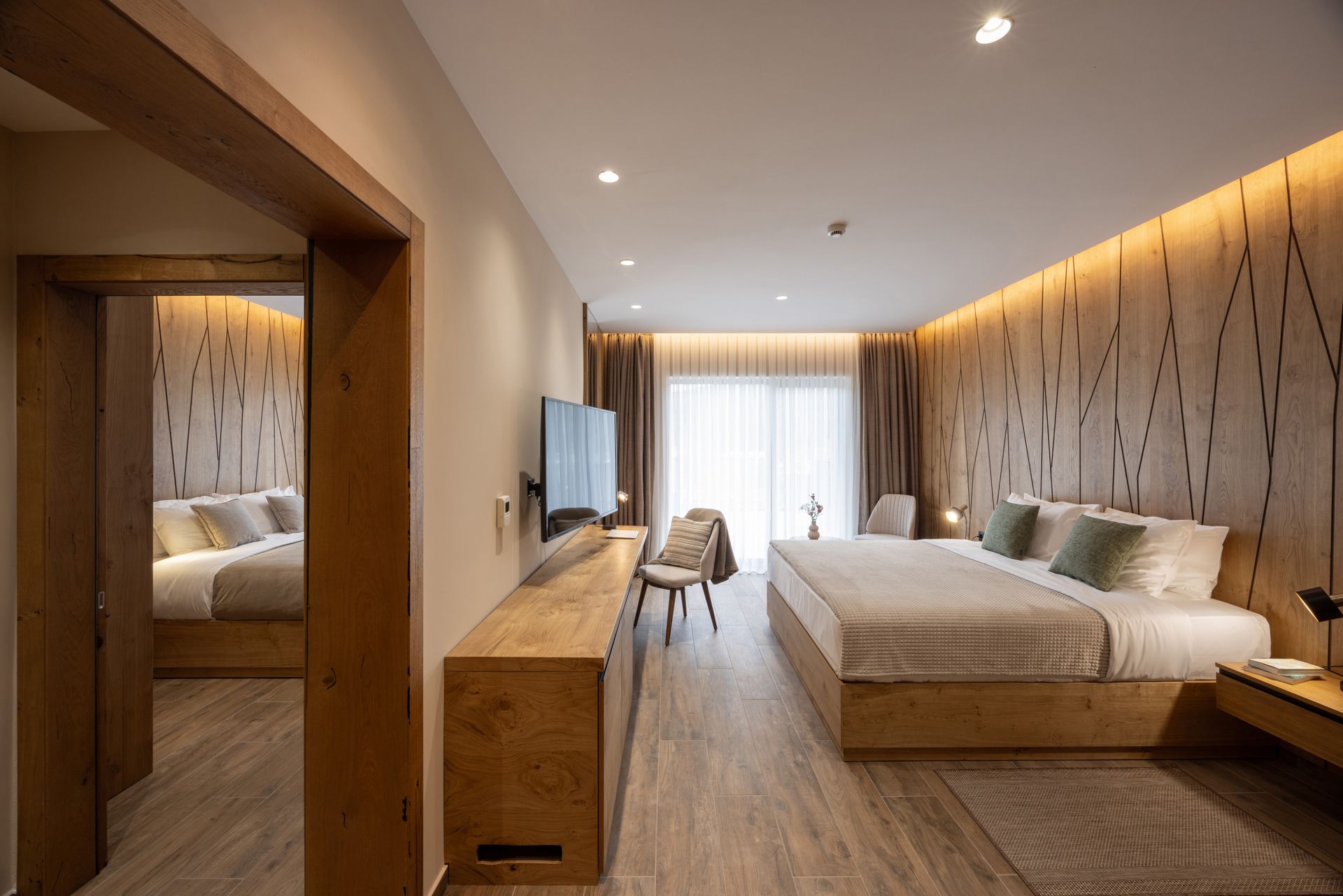 Modern hotel bedroom with wood-paneled walls, king bed, TV, and seating area near a bright window.