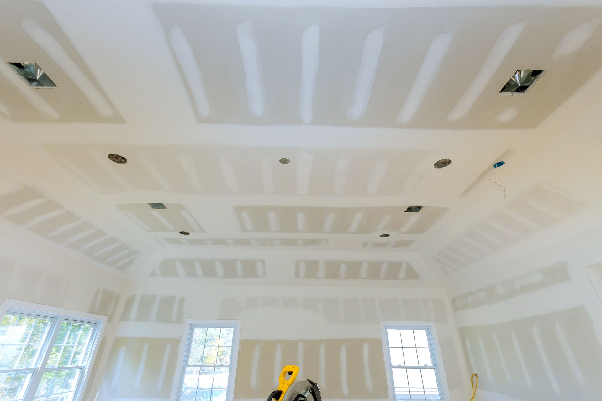 Bright white room under construction with drywall ceiling, recessed lights, and several windows.