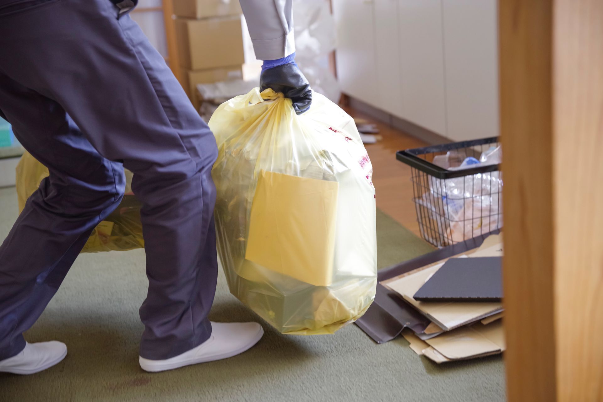 Person carrying a yellow trash bag past a pile of papers and a basket indoors