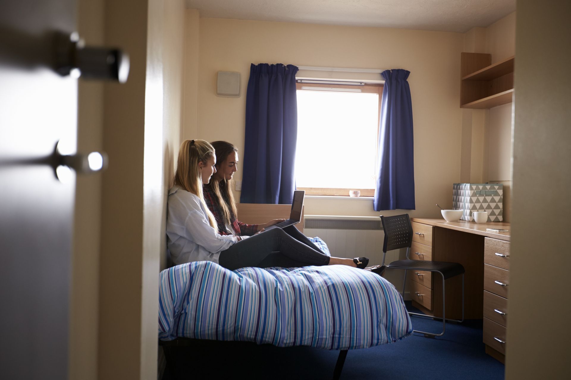 Two people sitting on a bed in a small bedroom, facing a bright window with blue curtains.