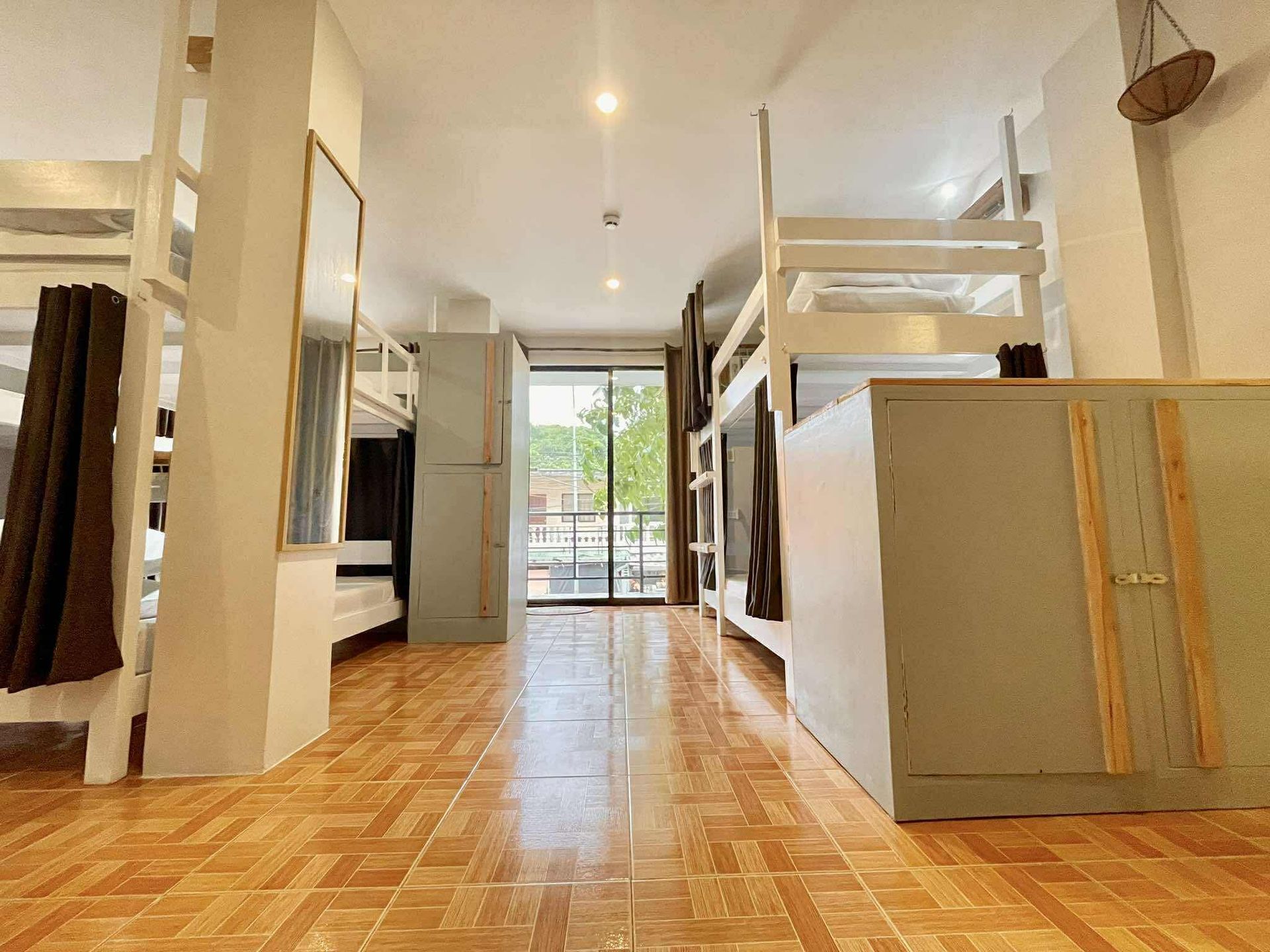 Bunk beds line a room with wood-look tile. Cabinets and a mirror stand between the beds. A balcony is at the end.