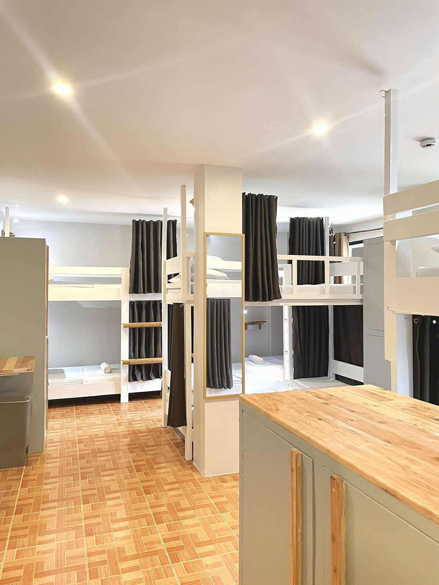 Hostel room with multiple white bunk beds, dark curtains, and wood cabinets.