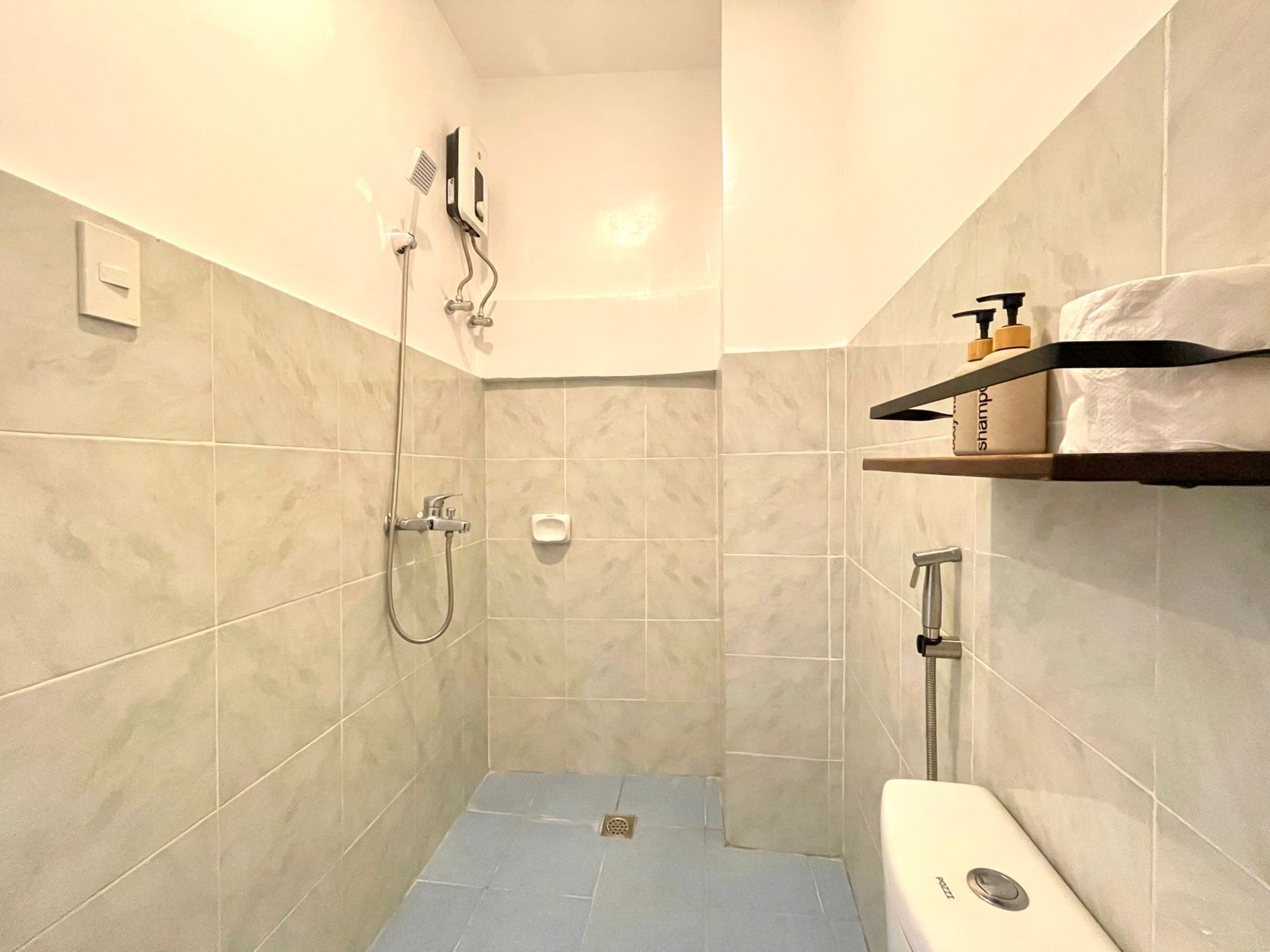 Small bathroom with tiled walls, shower, and toilet. A shelf holds toiletries and toilet paper.