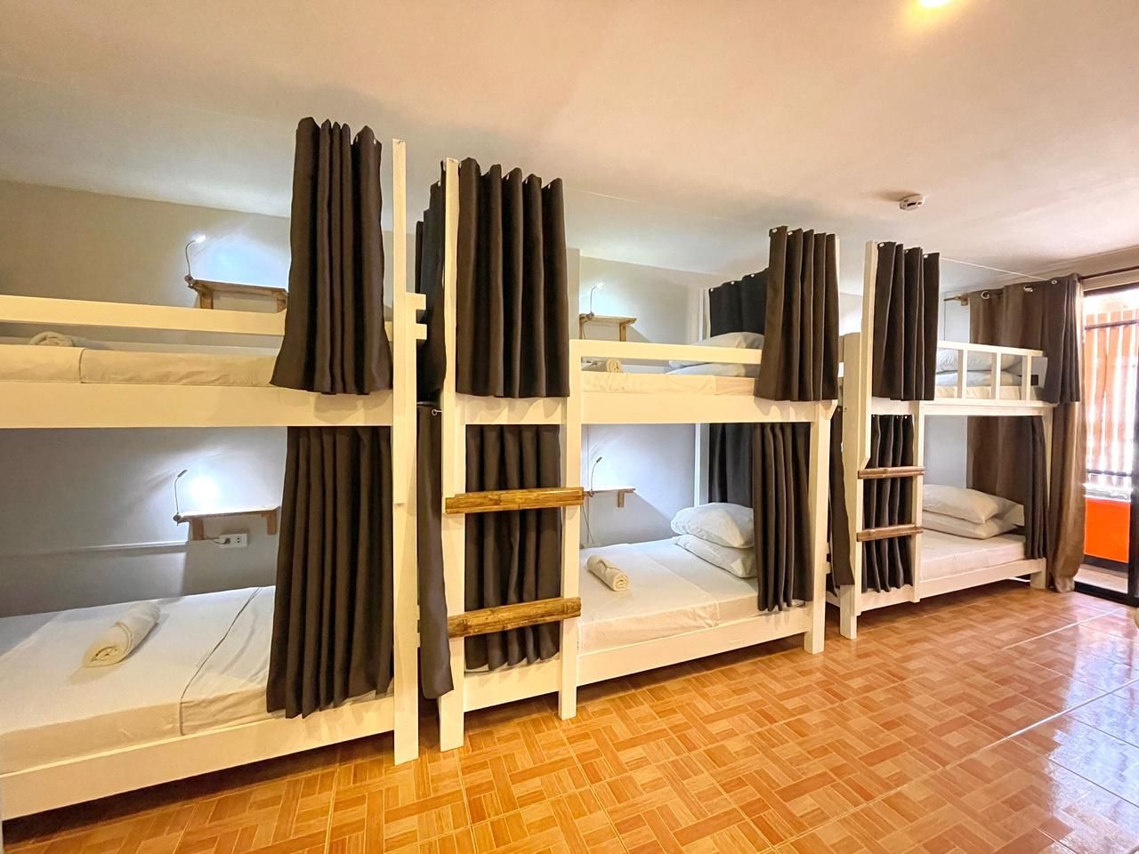 Hostel room with multiple white bunk beds, each with curtains. Wooden floors and walls, daytime.