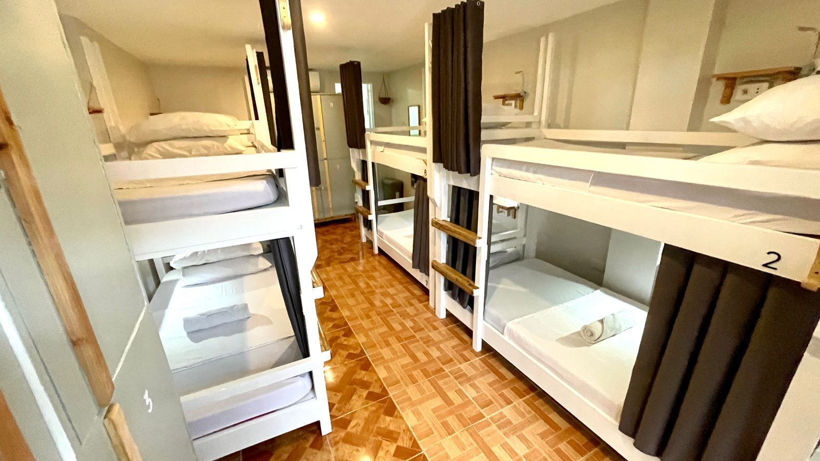 Dorm room with multiple bunk beds.  Each bed has a curtain for privacy. Wooden floor and white walls.