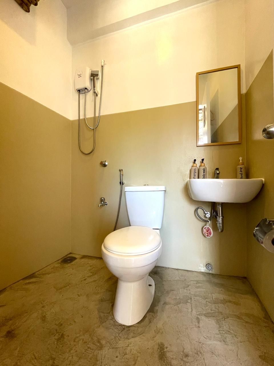 Bathroom with beige and white walls, toilet, sink, mirror, and showerhead on a concrete floor.