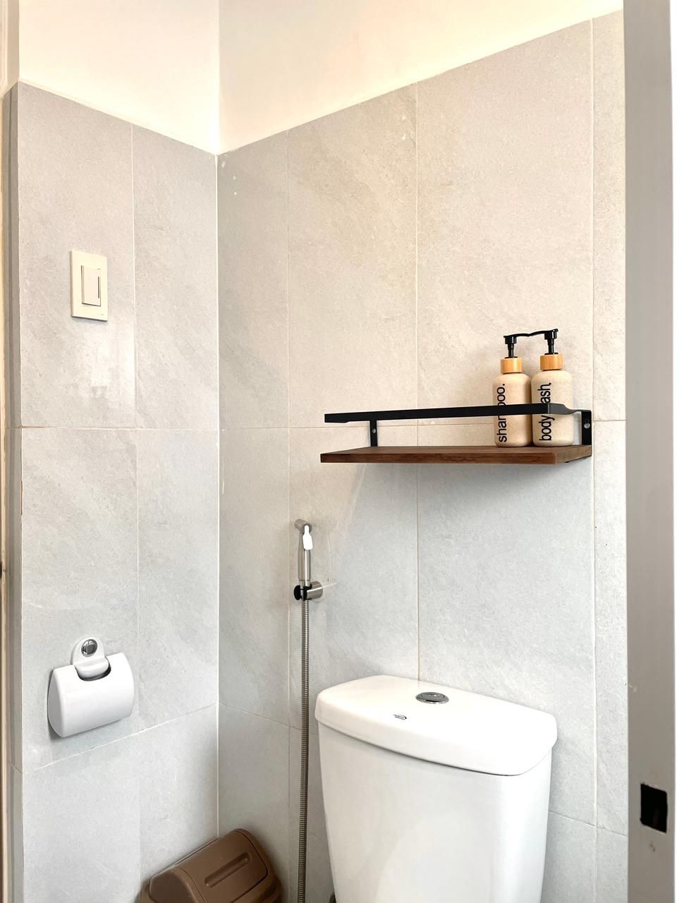 Bathroom corner with toilet, bidet, shelf with toiletries, and a roll of toilet paper on the wall.