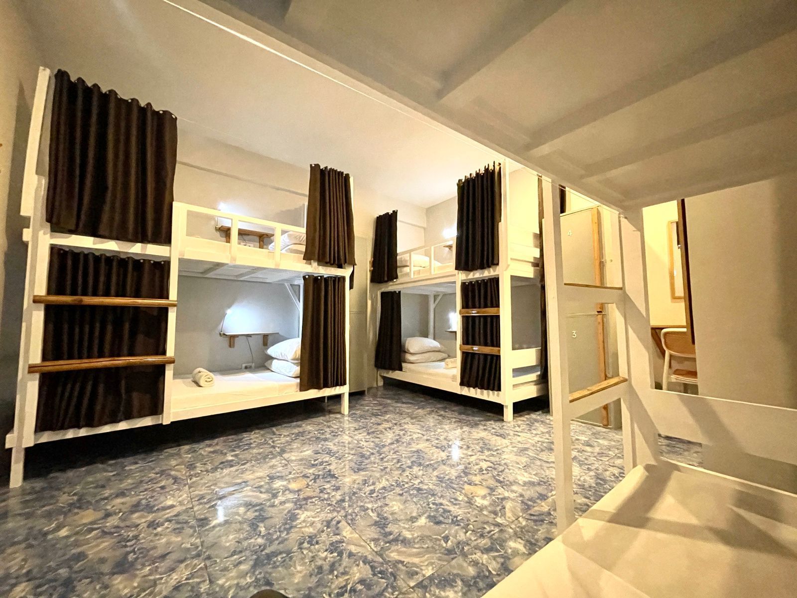 Hostel room with multiple white bunk beds and privacy curtains; blue tiled floor.