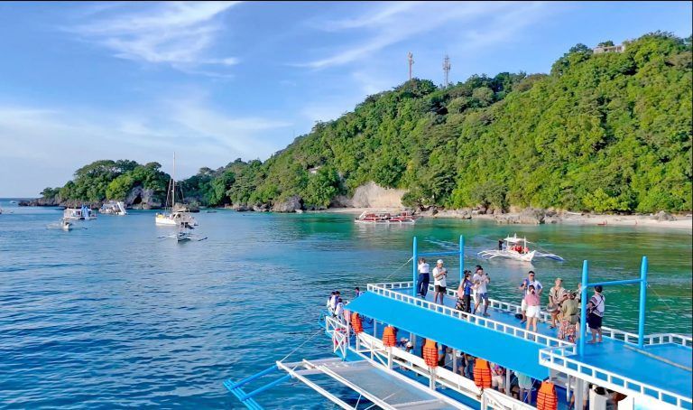 Blue boat with people on the water in a tropical cove, lush green hillside in the background.