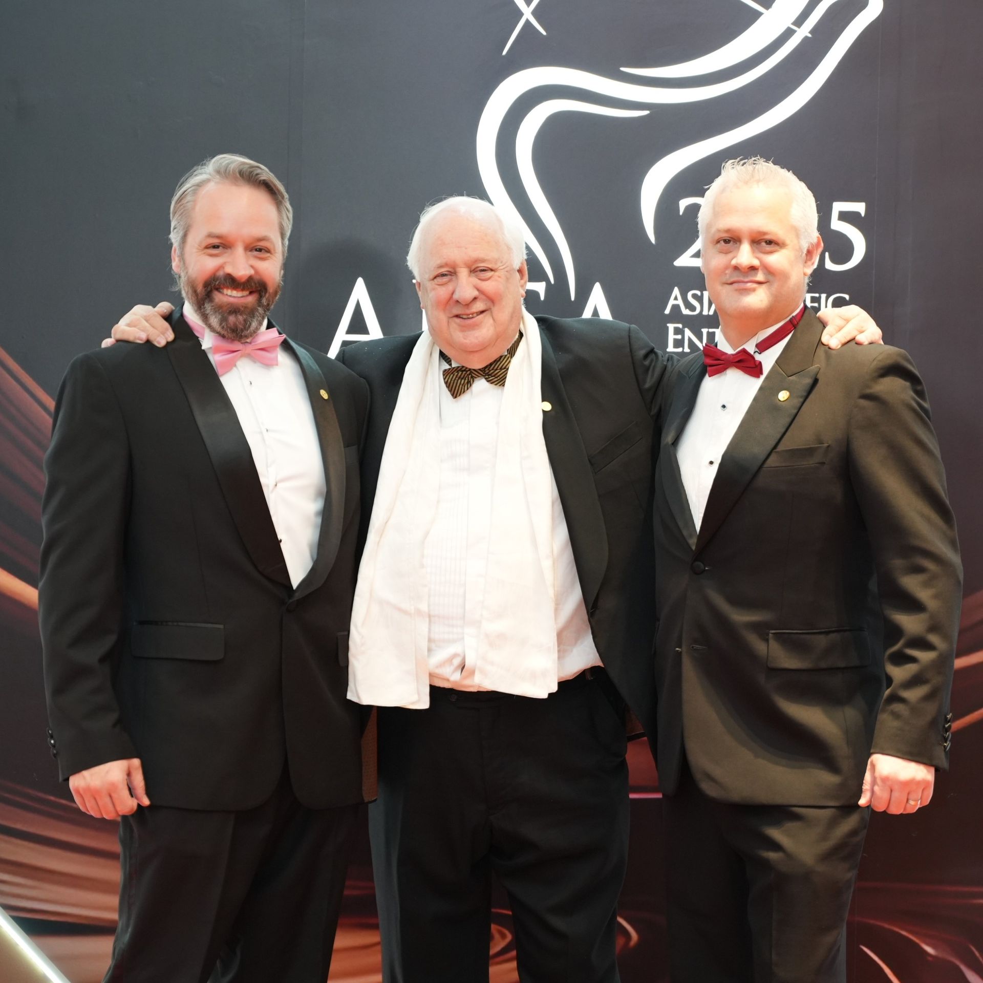 Three men in tuxedos, arms around each other, pose in front of a backdrop with text and a logo.