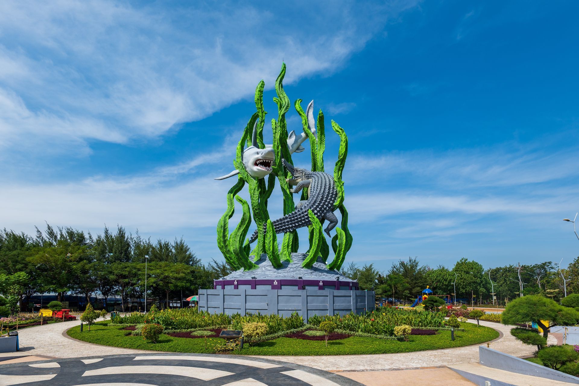 Monument featuring a shark and crocodile amidst green seaweed, set against a blue sky, in a park.