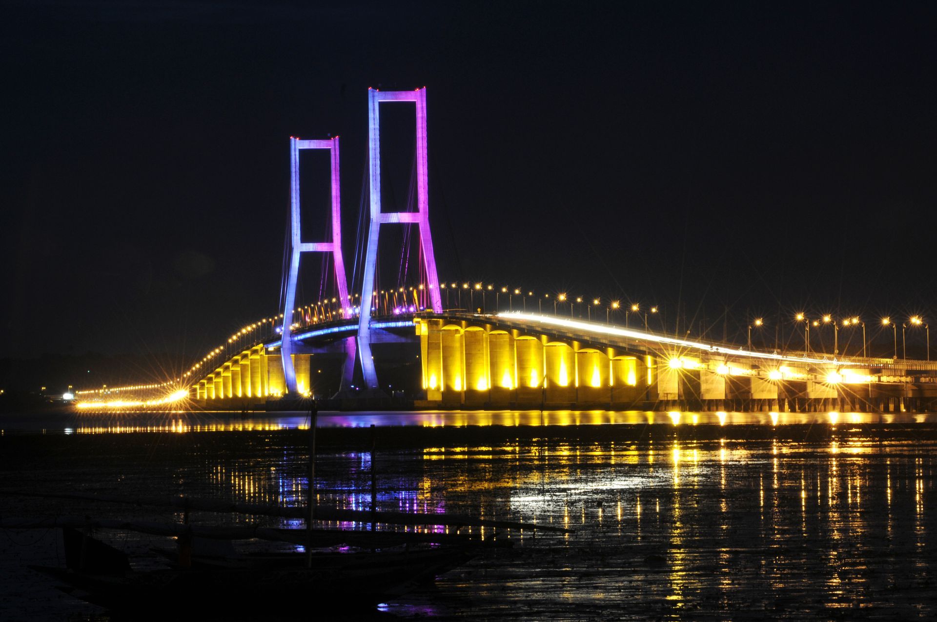 A brightly lit cable-stayed bridge at night, with purple and gold lights reflecting in the water.