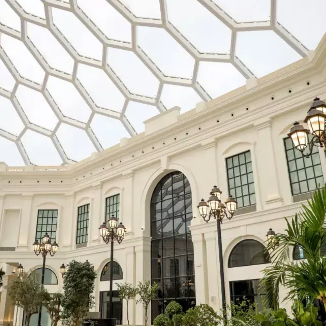 Interior of a white building with a hexagonal glass ceiling; ornate street lamps and potted plants.
