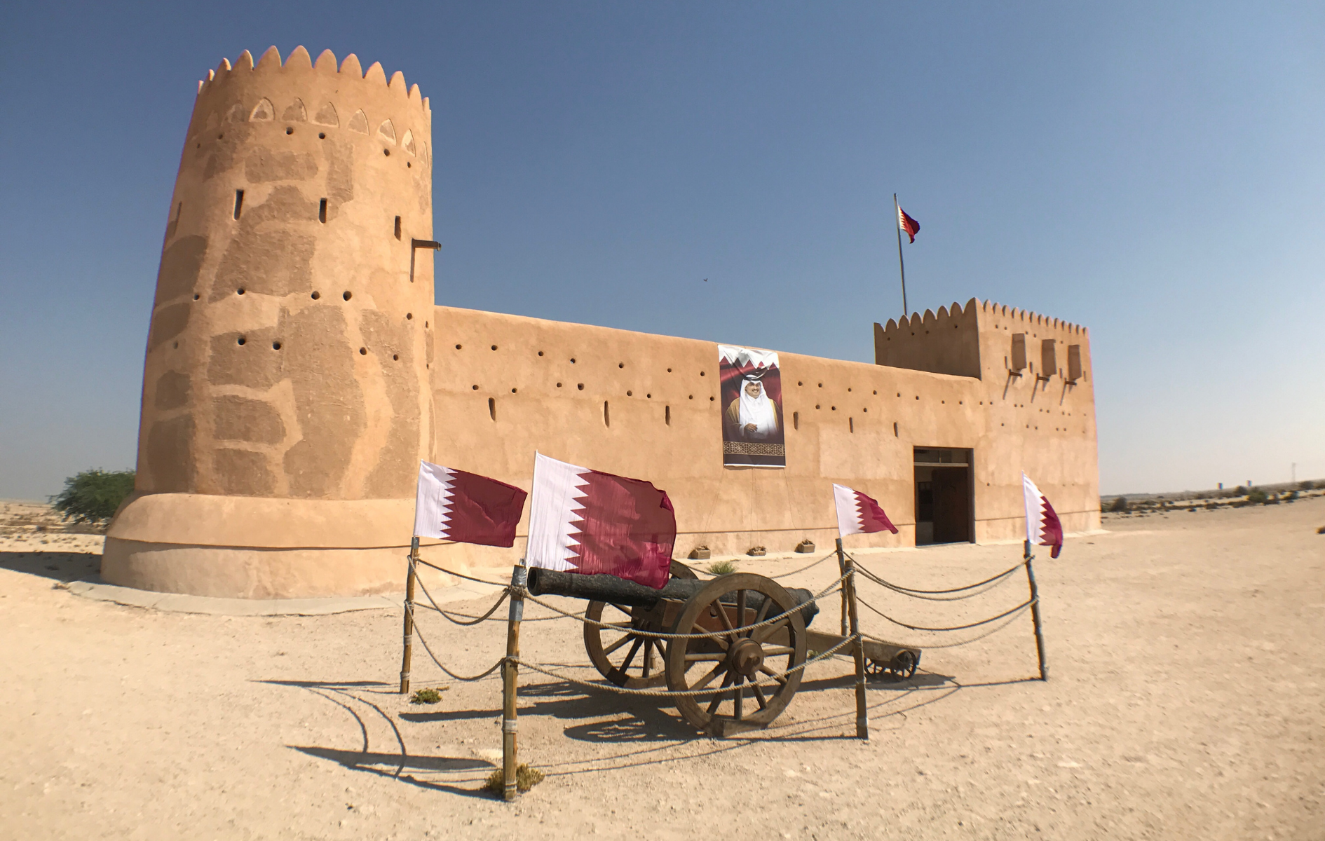 Sand-colored fort with round tower and flags. Cannon in foreground, desert setting under a blue sky.