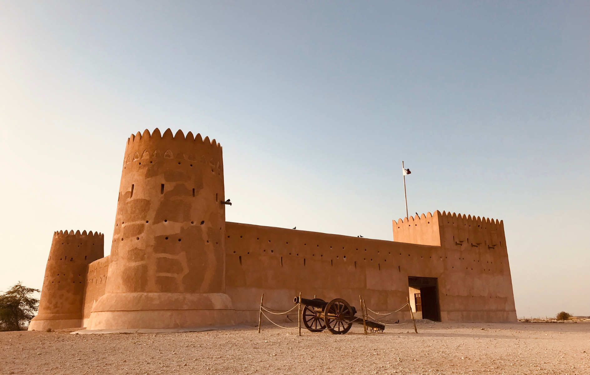 Desert fort with three crenellated towers and a cannon in front; clear sky.