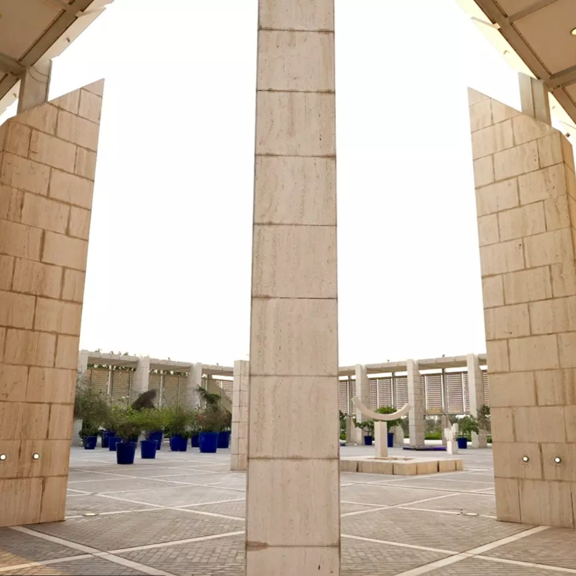 An outdoor courtyard with light stone columns, trees in blue pots, and a fountain.