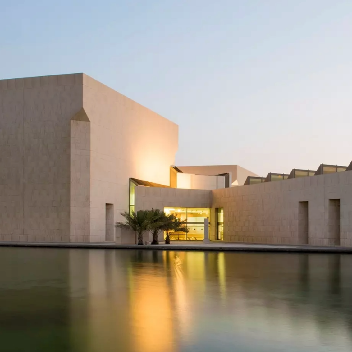 Modern building with a rectangular design reflected in calm water. Entryway glows with interior light.