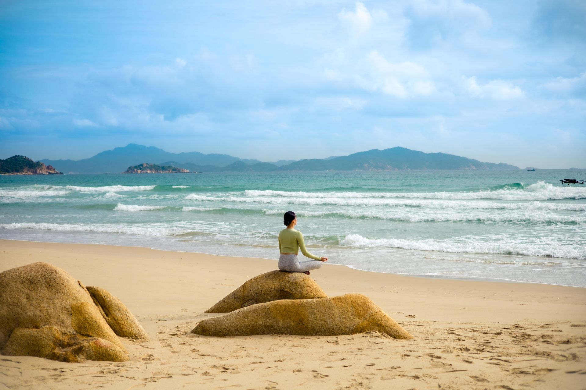 Person meditating on a rock at a beach, ocean and mountains in the background.