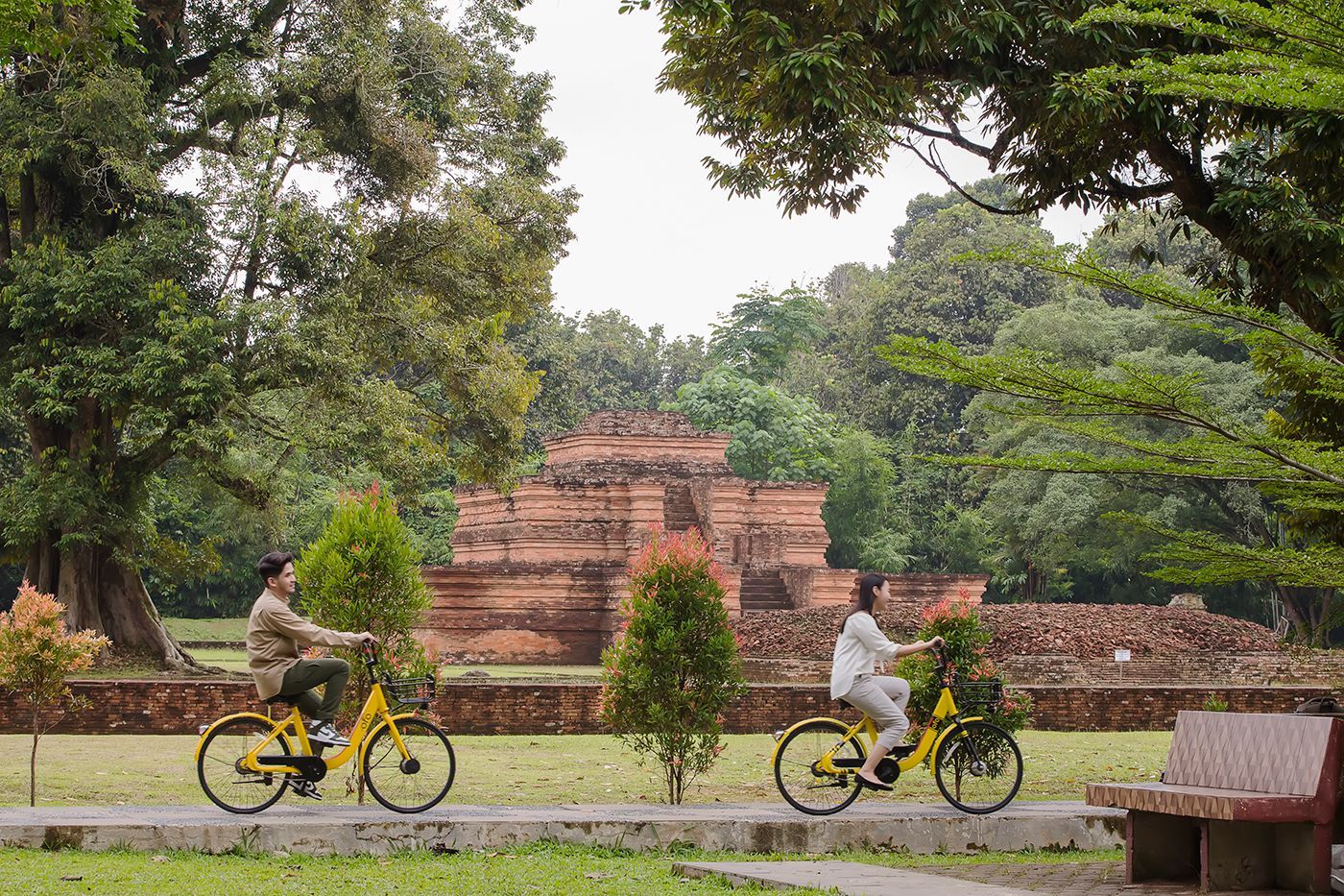Two people ride yellow bikes in a park, historic brick structure in the background.