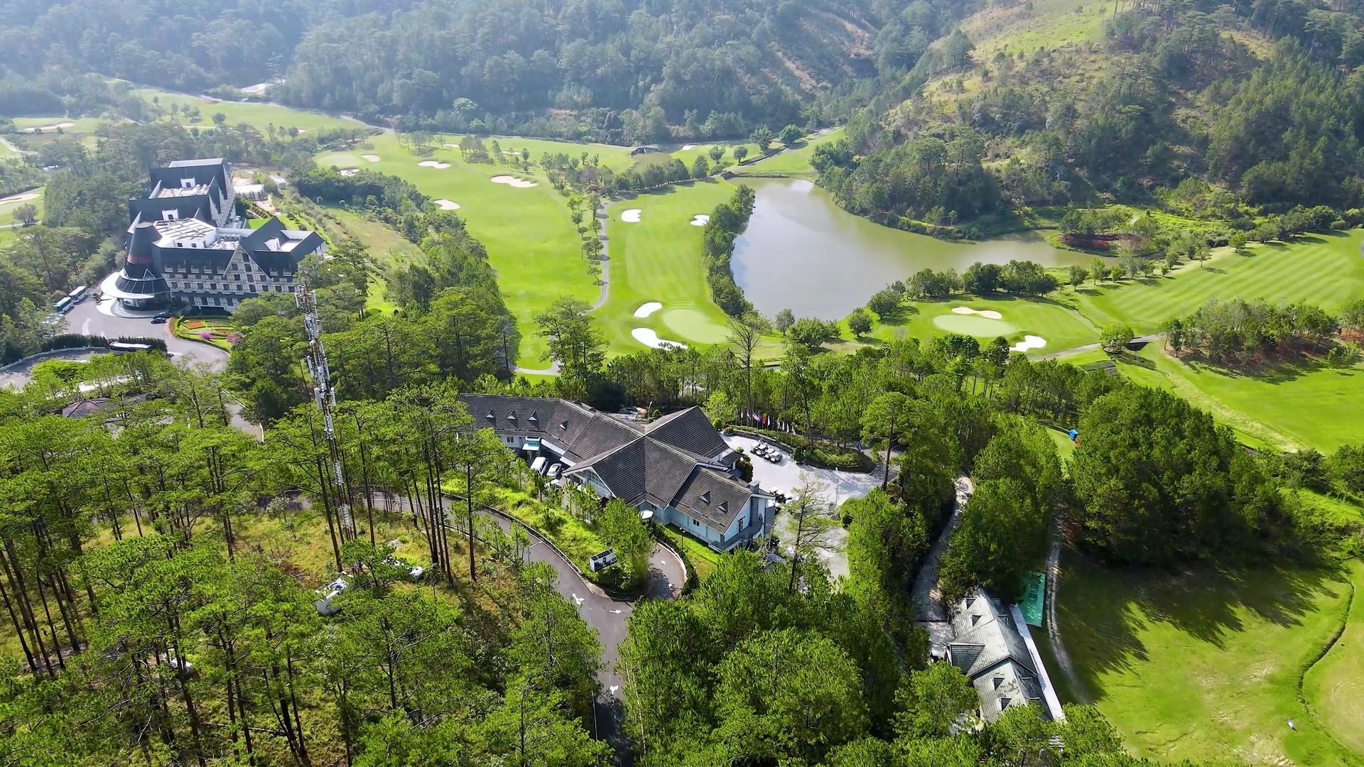 Aerial view of a golf course with a lake and buildings surrounded by green trees and hills.