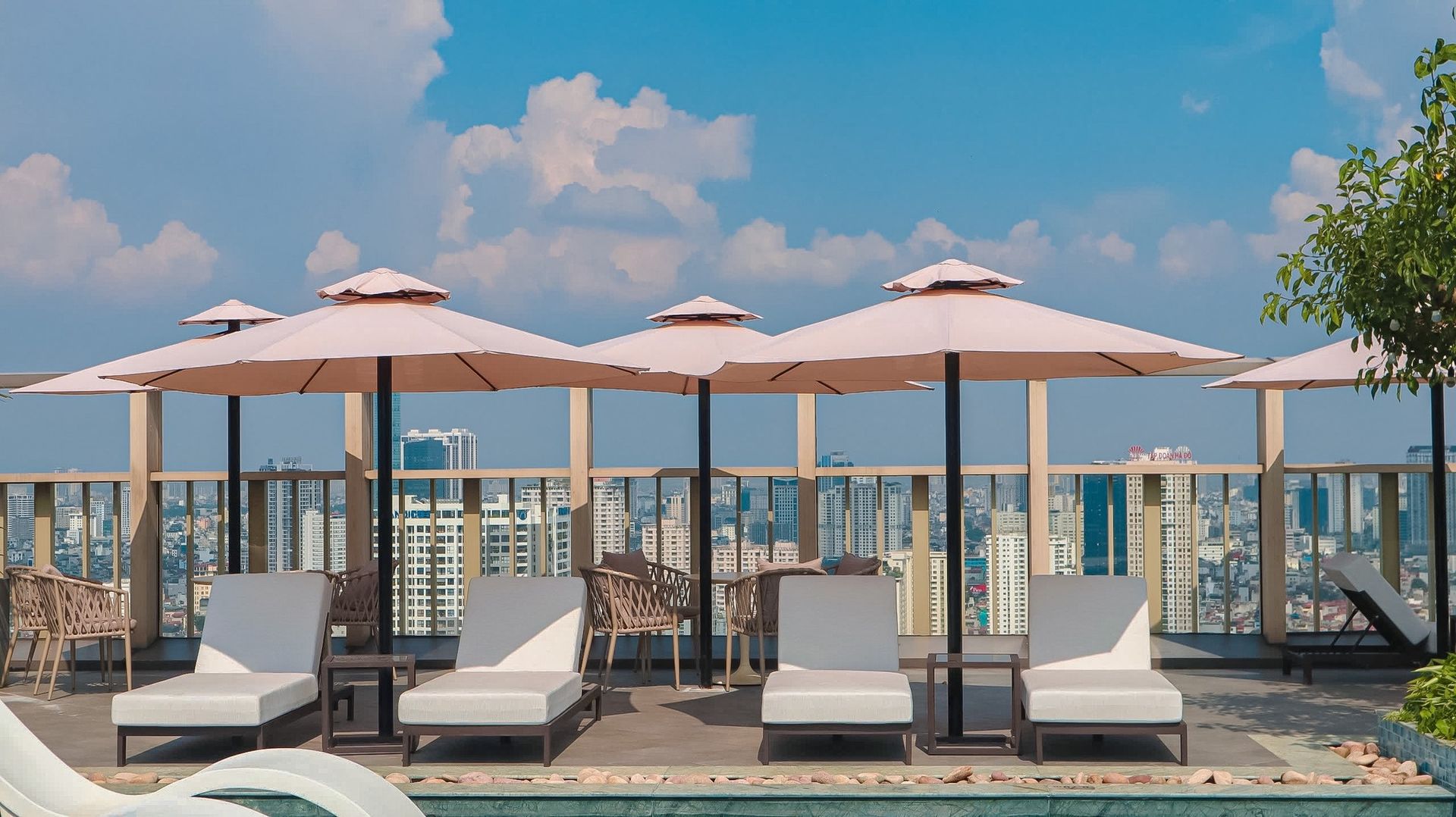 Rooftop pool with lounge chairs, umbrellas, and city view under a blue sky with clouds.
