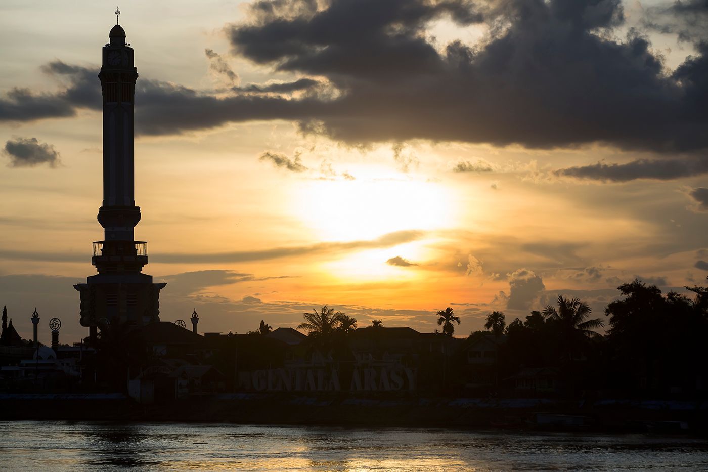 Silhouette of a tall minaret and buildings against a sunset sky over water.