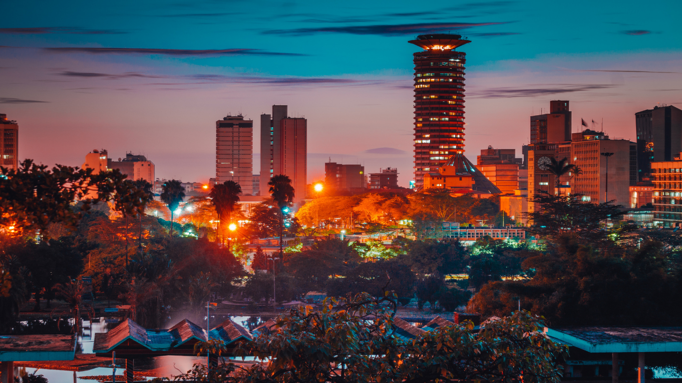 City skyline at dusk with lit buildings, orange and blue hues in the sky, trees in the foreground.