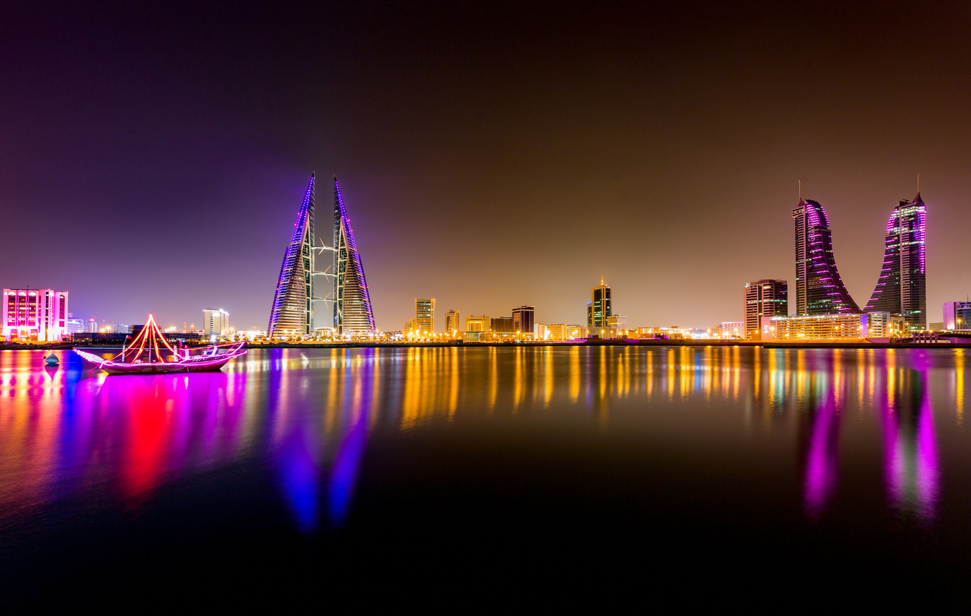 Nighttime cityscape reflecting in water; Bahrain, with lit skyscrapers and boat.