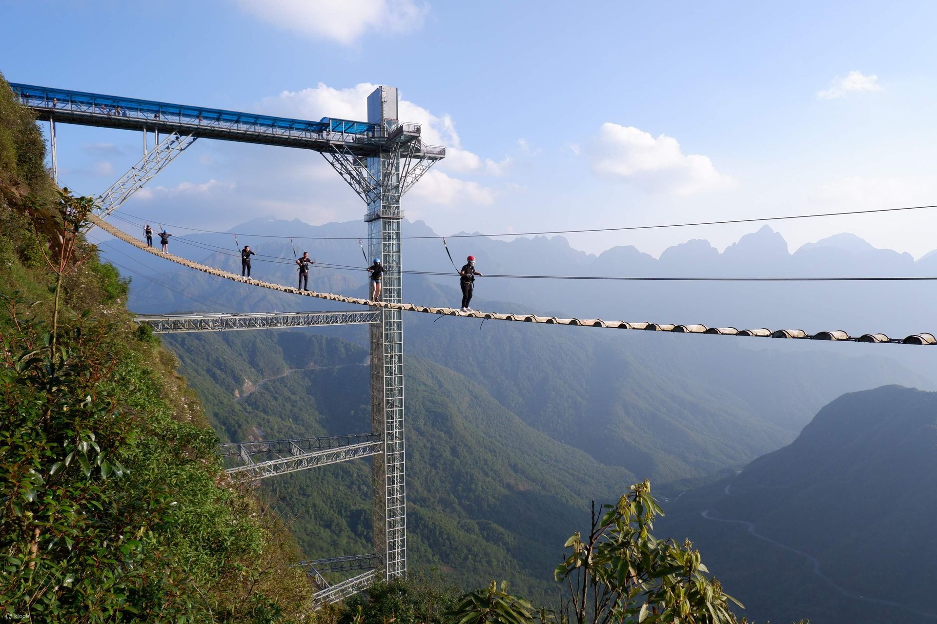 People walk on a high, narrow bridge over a deep valley. The bridge is supported by cables and towers.