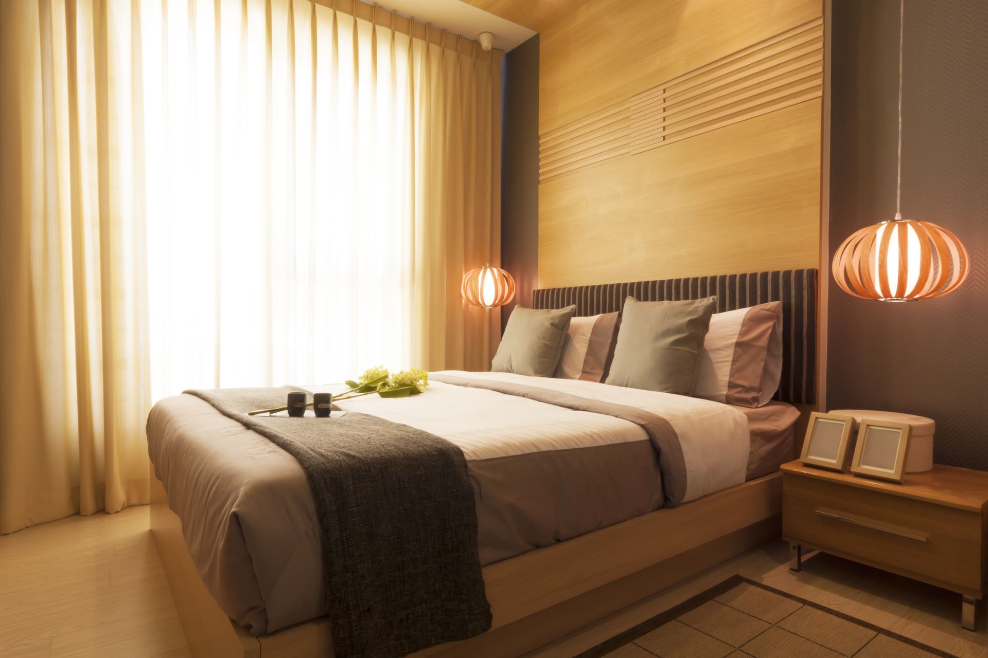 Cozy bedroom with a bed, nightstand, and window with curtains; neutral tones and hanging lamps.