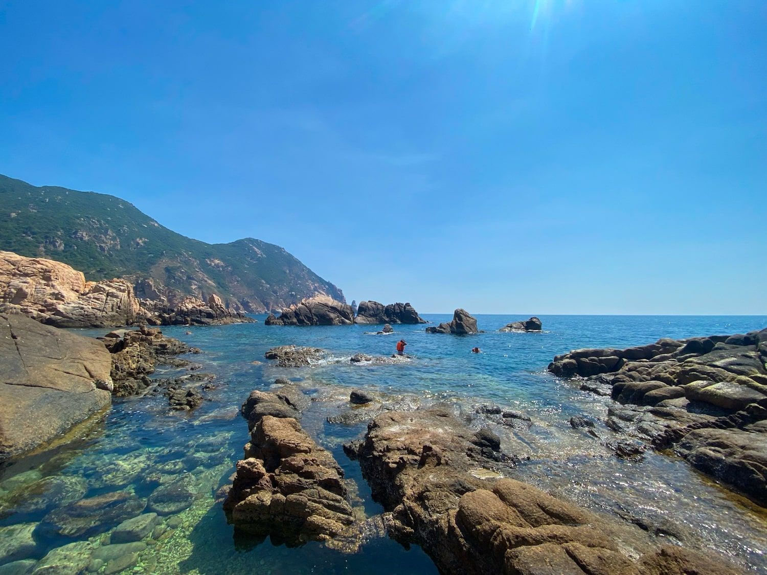 Rocky coastline with clear blue water and mountain under a bright blue sky.