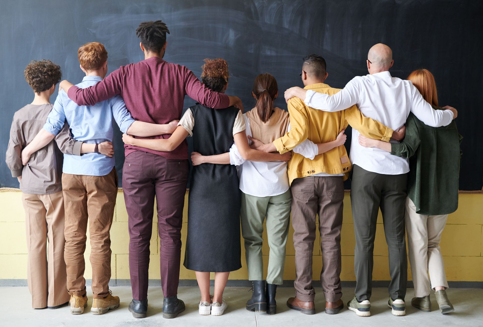 Group of people standing arm-in-arm in a line, facing a wall in a bright room.