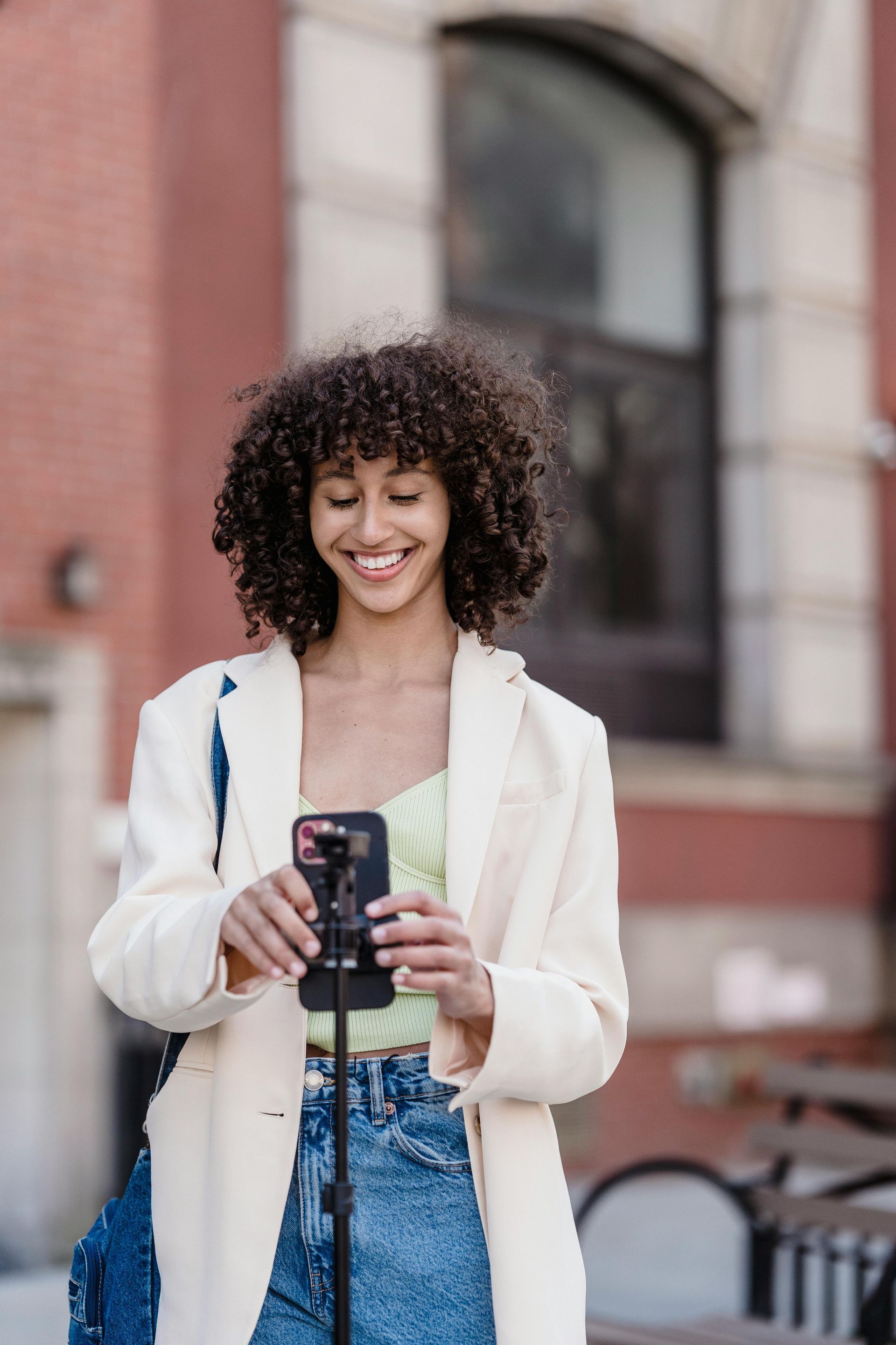 Woman smiles while holding a phone attached to a tripod outdoors. She wears a white blazer and jeans. Red brick building in background.
