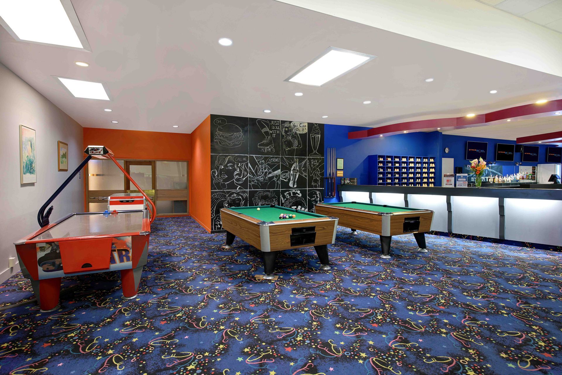Colorful indoor arcade with pool tables, neon-lit walls, and blue patterned carpet
