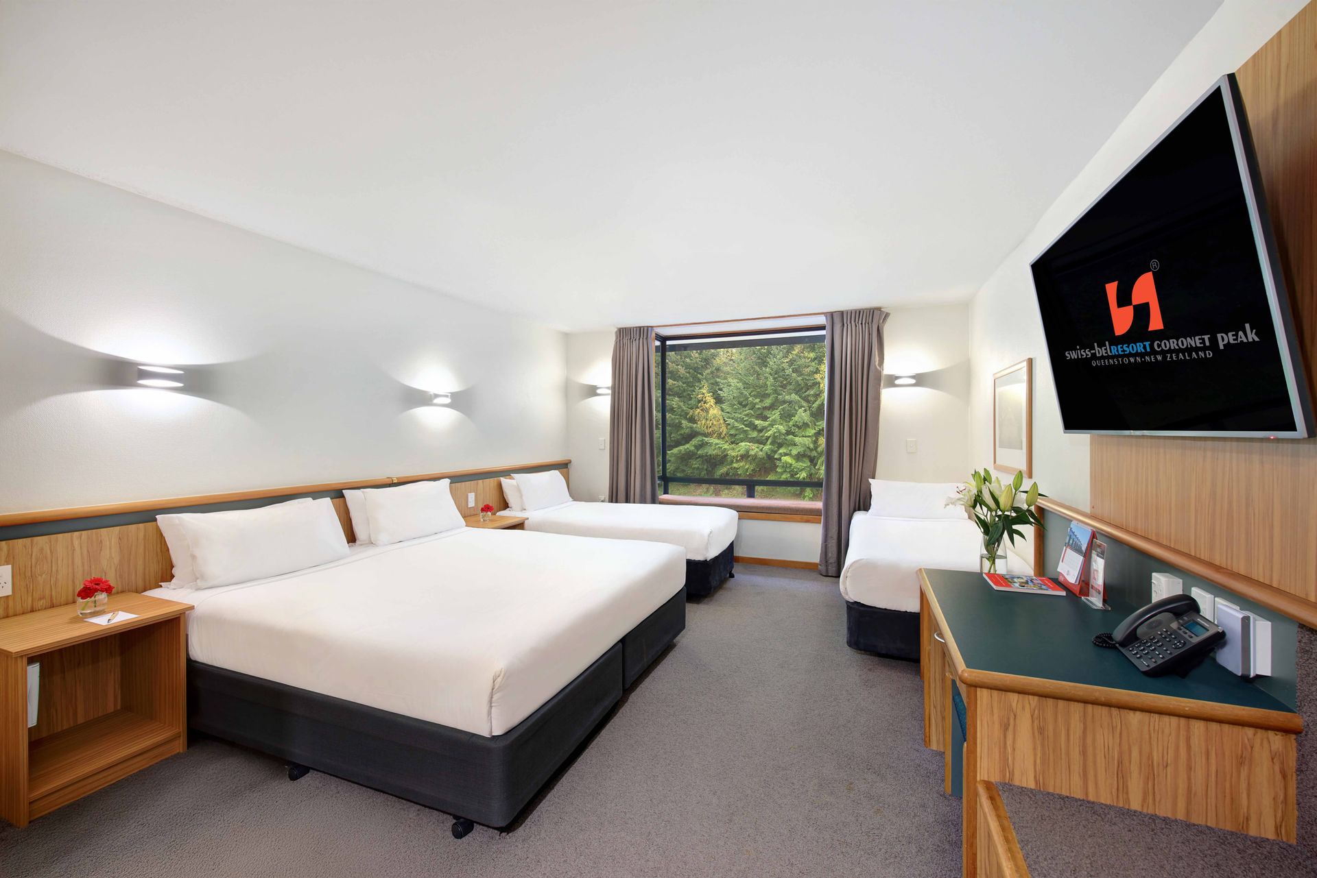 Modern hotel room with two beds, wall-mounted TV, desk, and window seat under skylight.