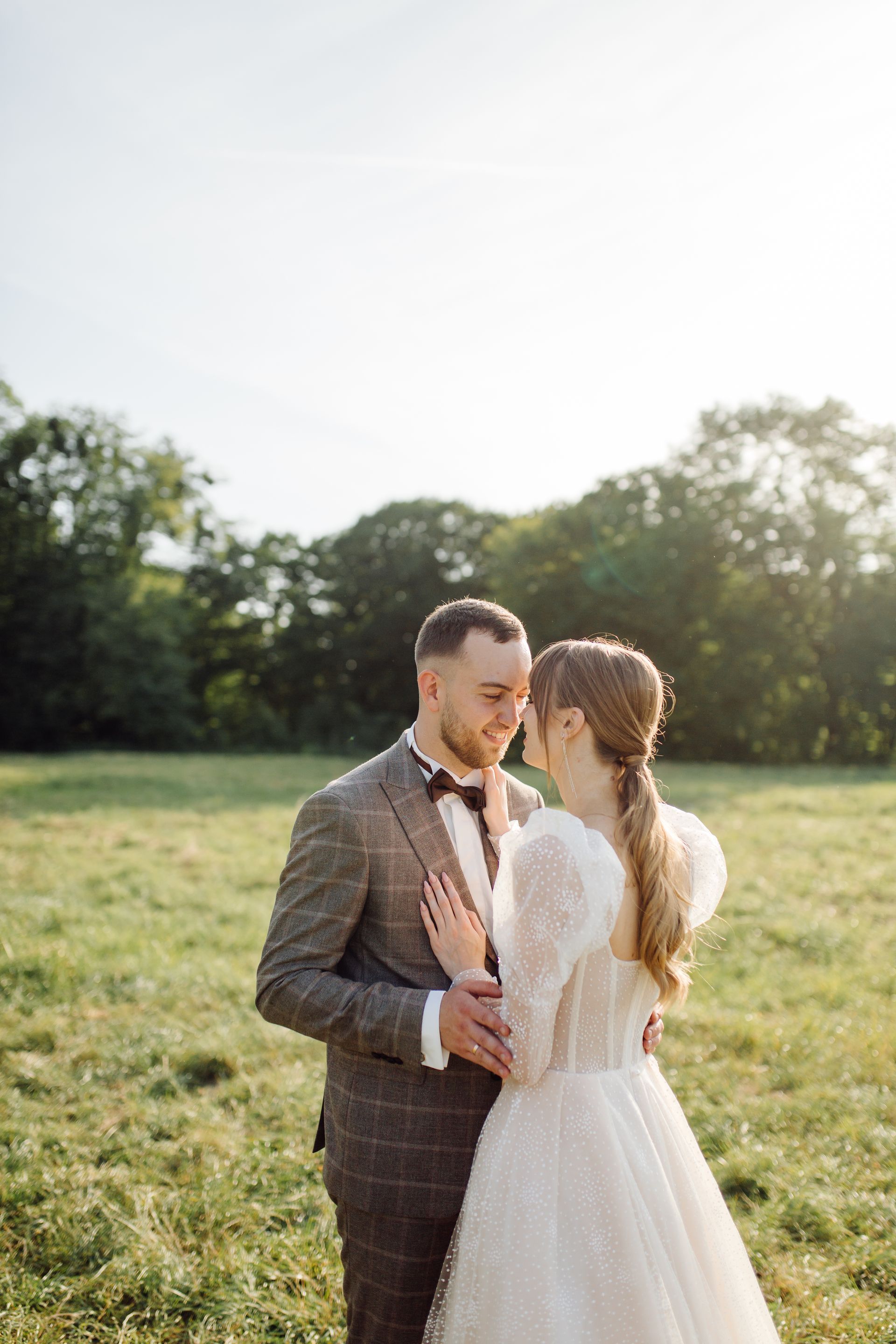 Couple embraces in a sunny field, the bride in a white dress, the groom in a checkered suit.