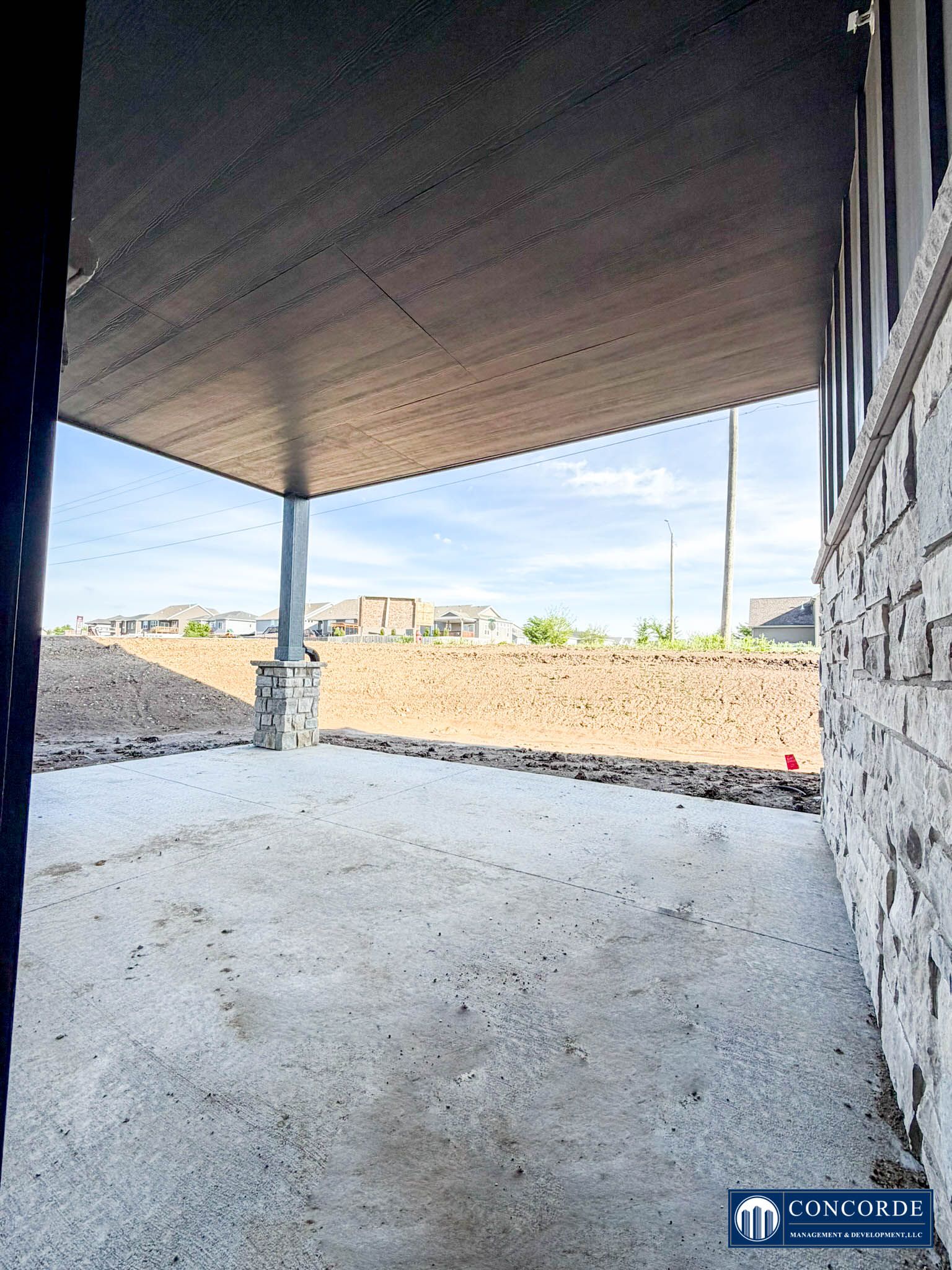 Covered porch with concrete floor and ceiling, overlooking a dirt lot and neighboring buildings under a clear sky.