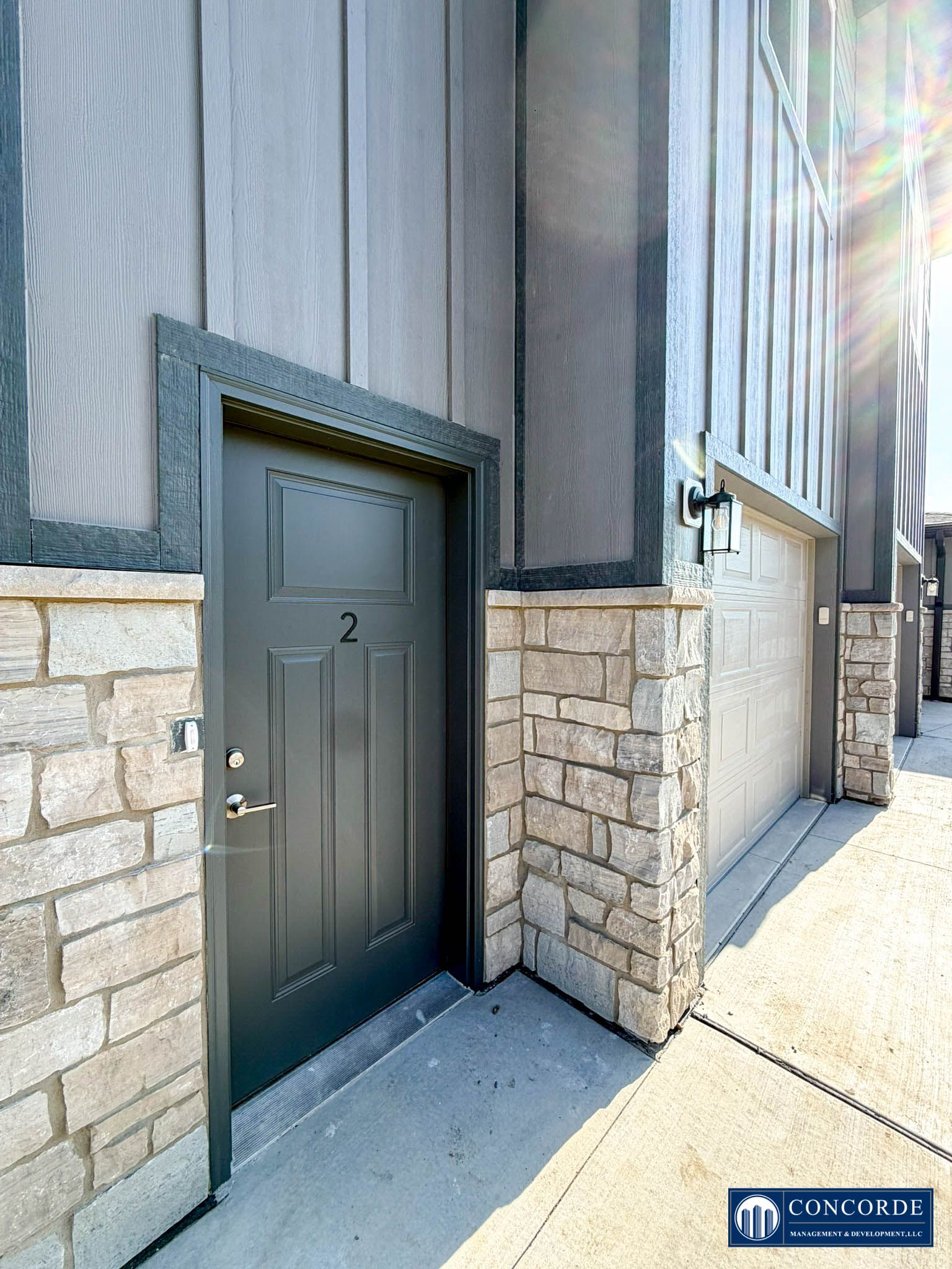 Black door in a stone-and-gray building exterior beside a garage door on a sidewalk
