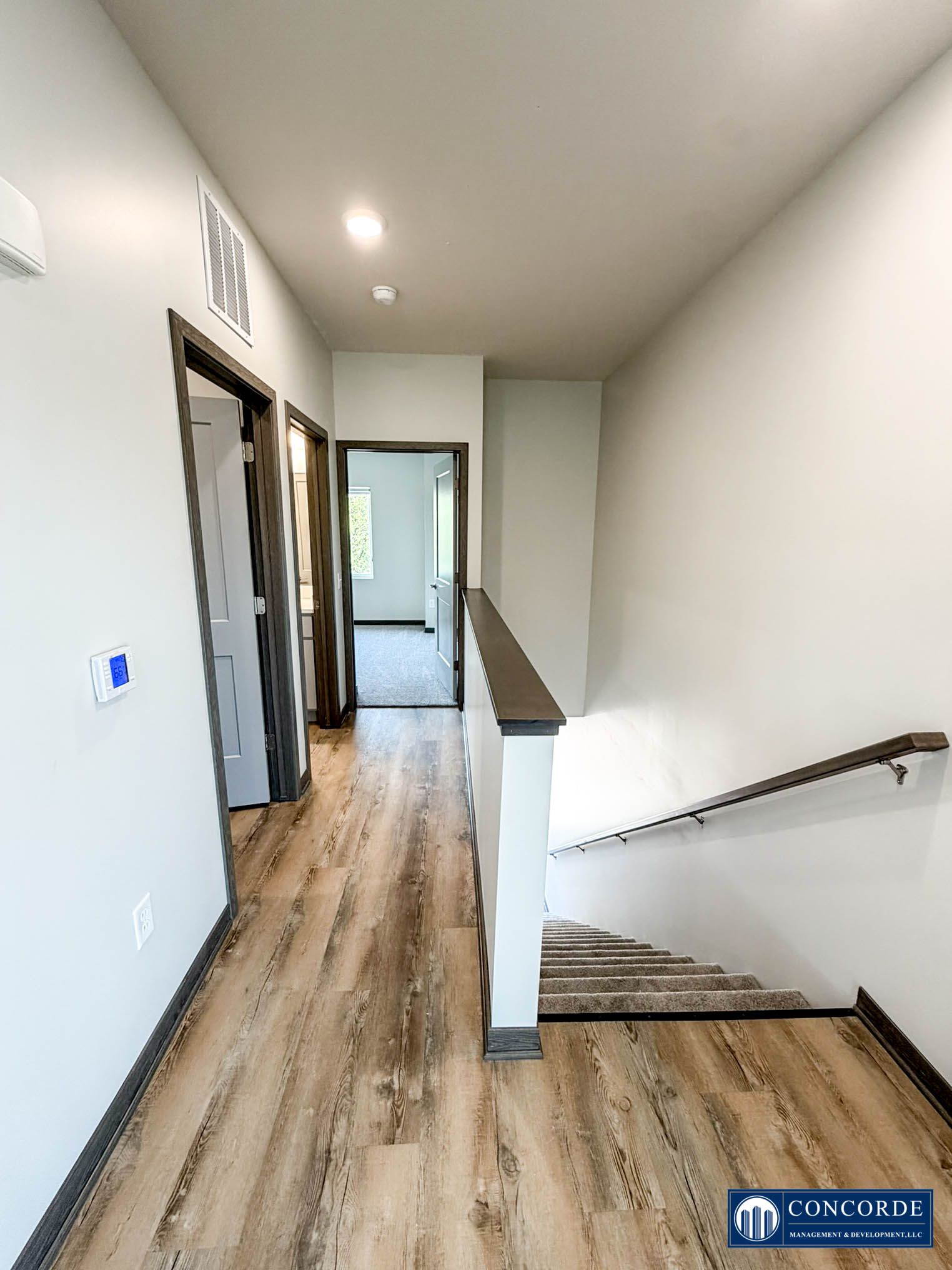 Narrow hallway with wood floors, white walls, and staircase railing leading to a door at the far end