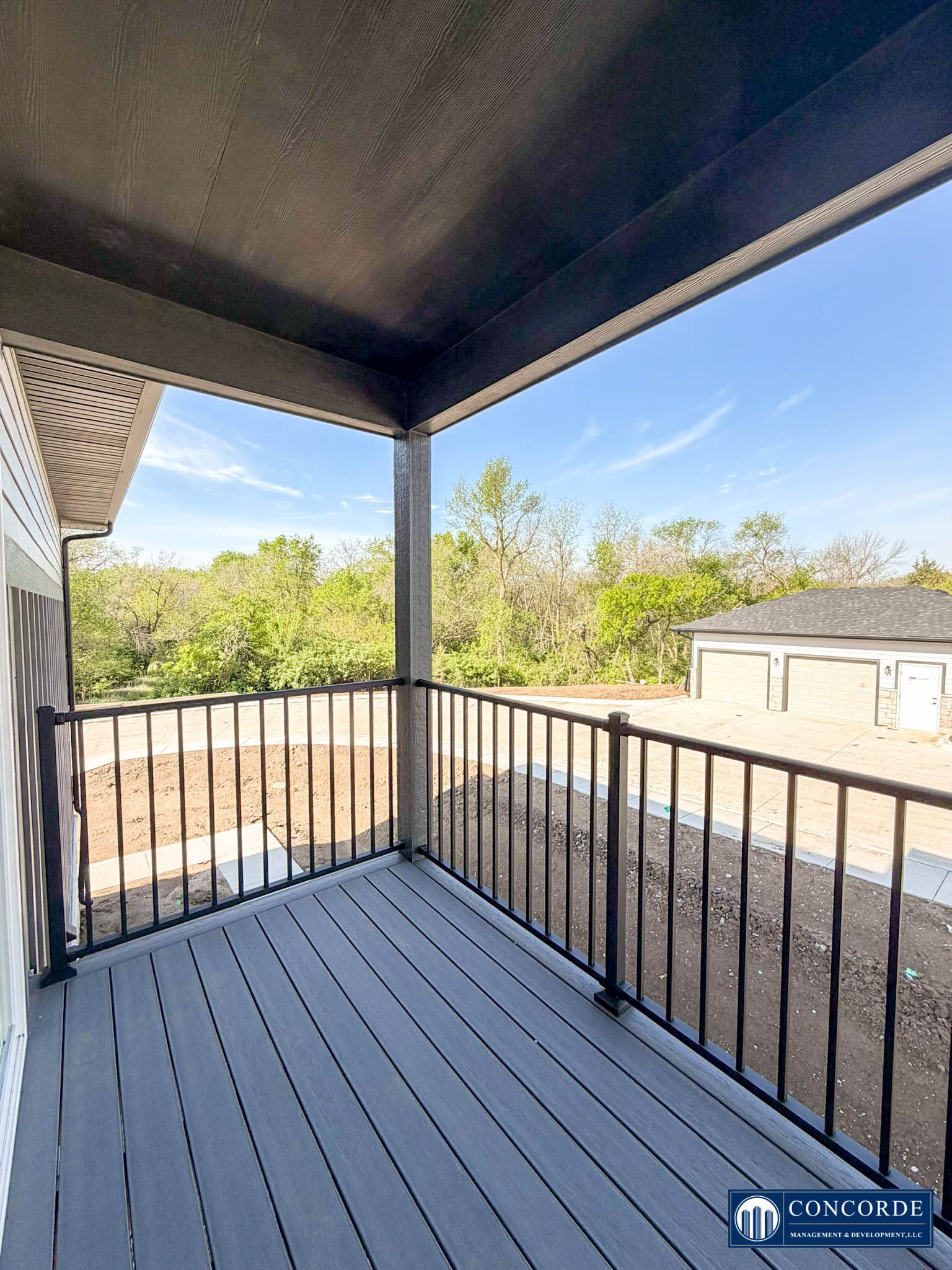 Covered balcony with black railing, gray floor, and view of trees and neighboring buildings.