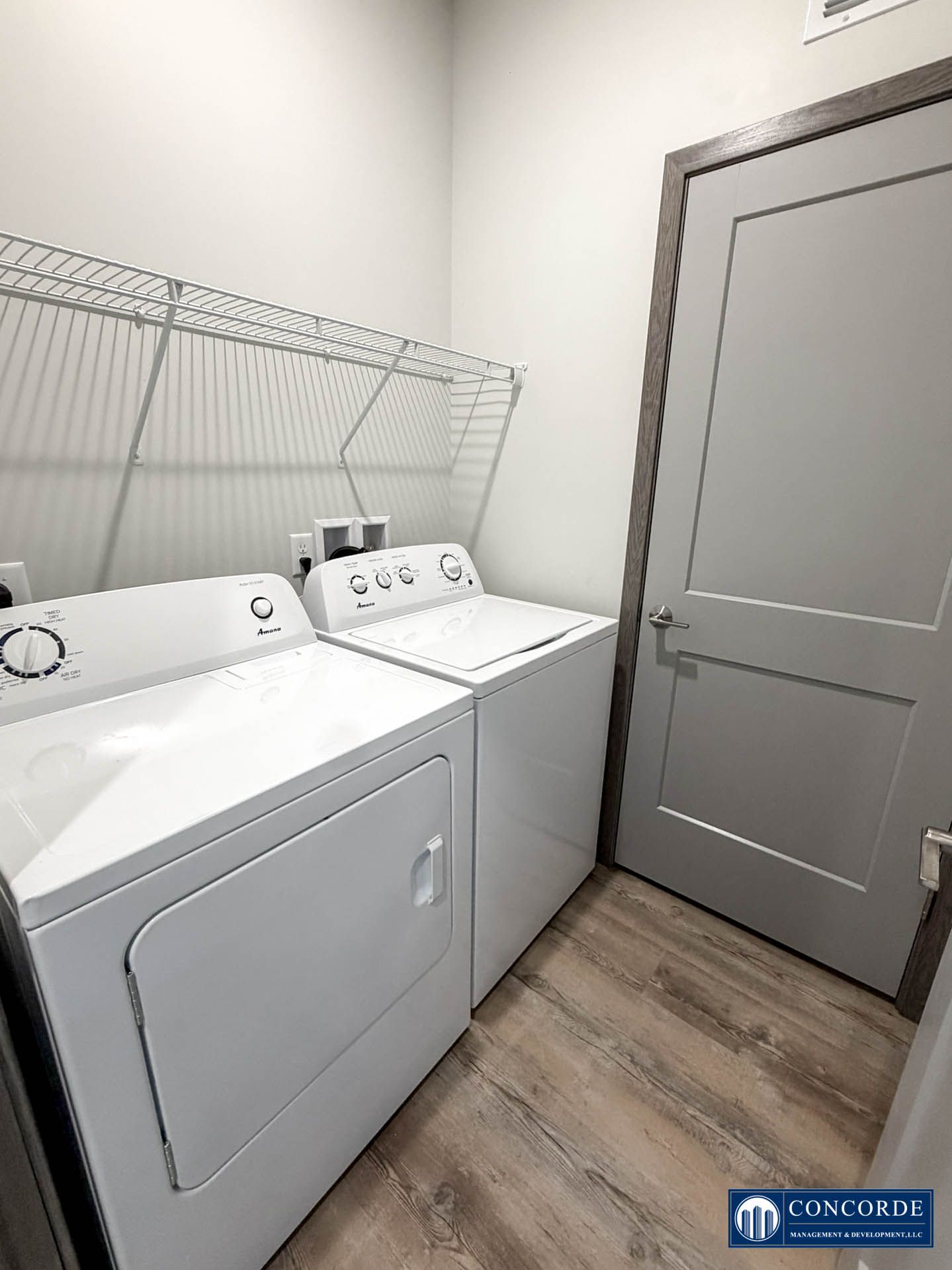 Laundry room with white washer and dryer, wire shelf, and closed gray door.