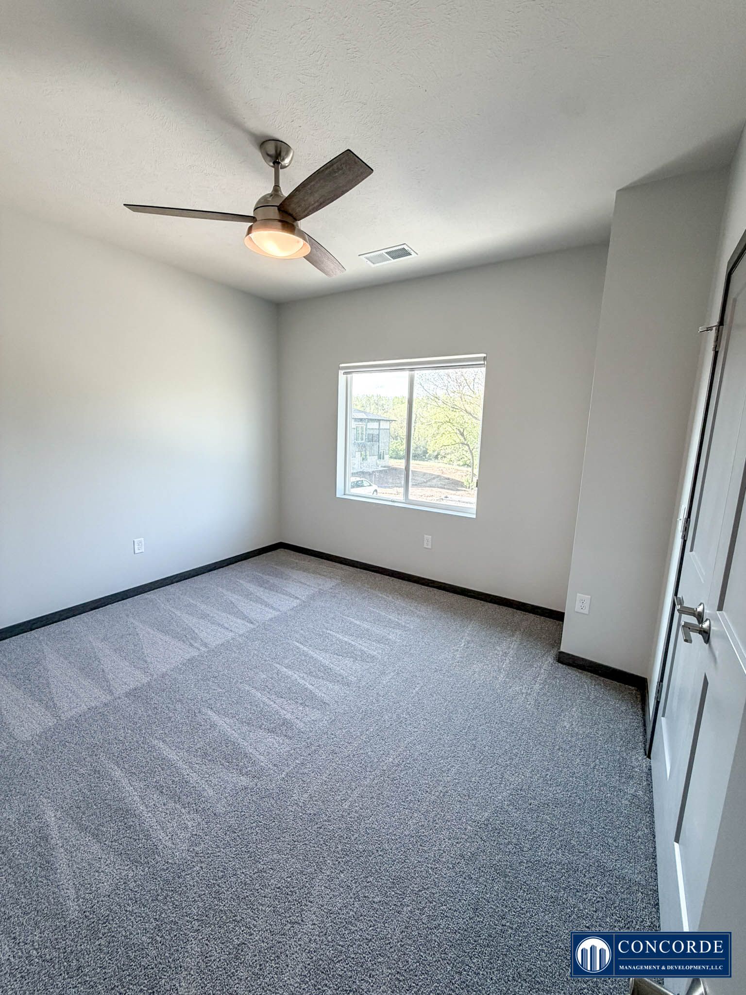 Empty carpeted bedroom with white walls, window, closet doors, and a ceiling fan with light.