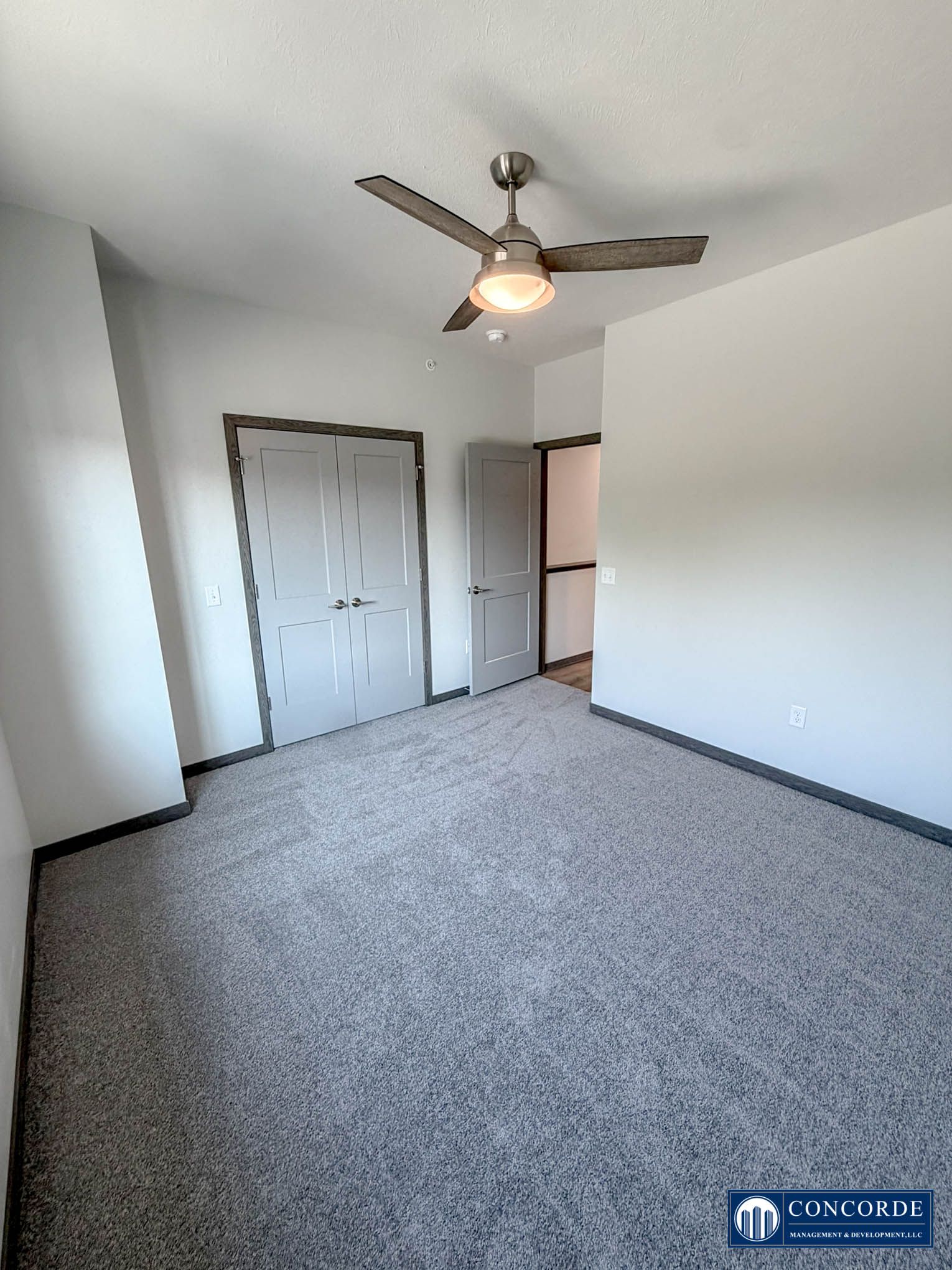 Empty bedroom with gray carpet, ceiling fan, closed double closet doors, and an open doorway.