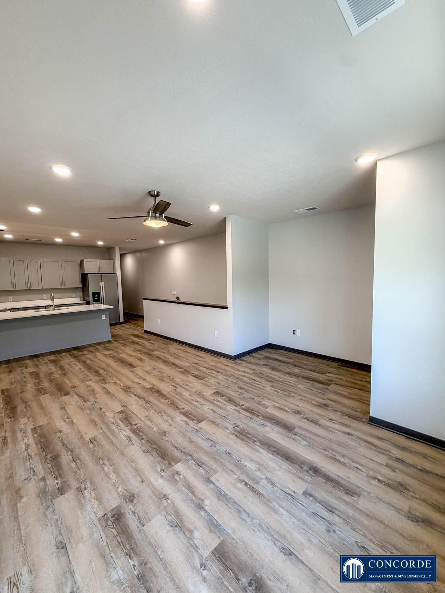 Spacious empty living room with wood floors, white walls, ceiling fan, and open kitchen area.