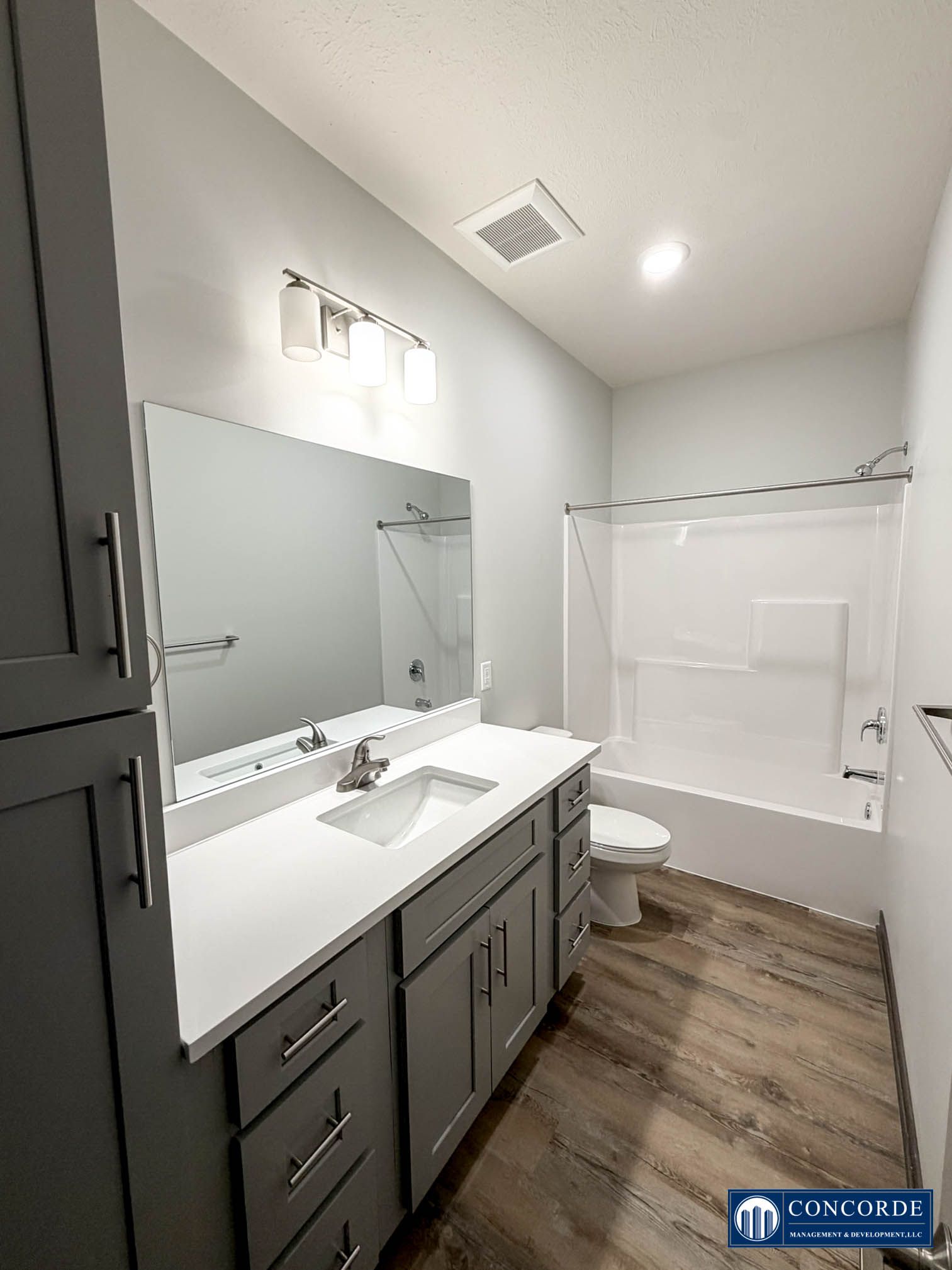 Bright bathroom with gray vanity, large mirror, white tub-shower combo, and wood-look flooring.