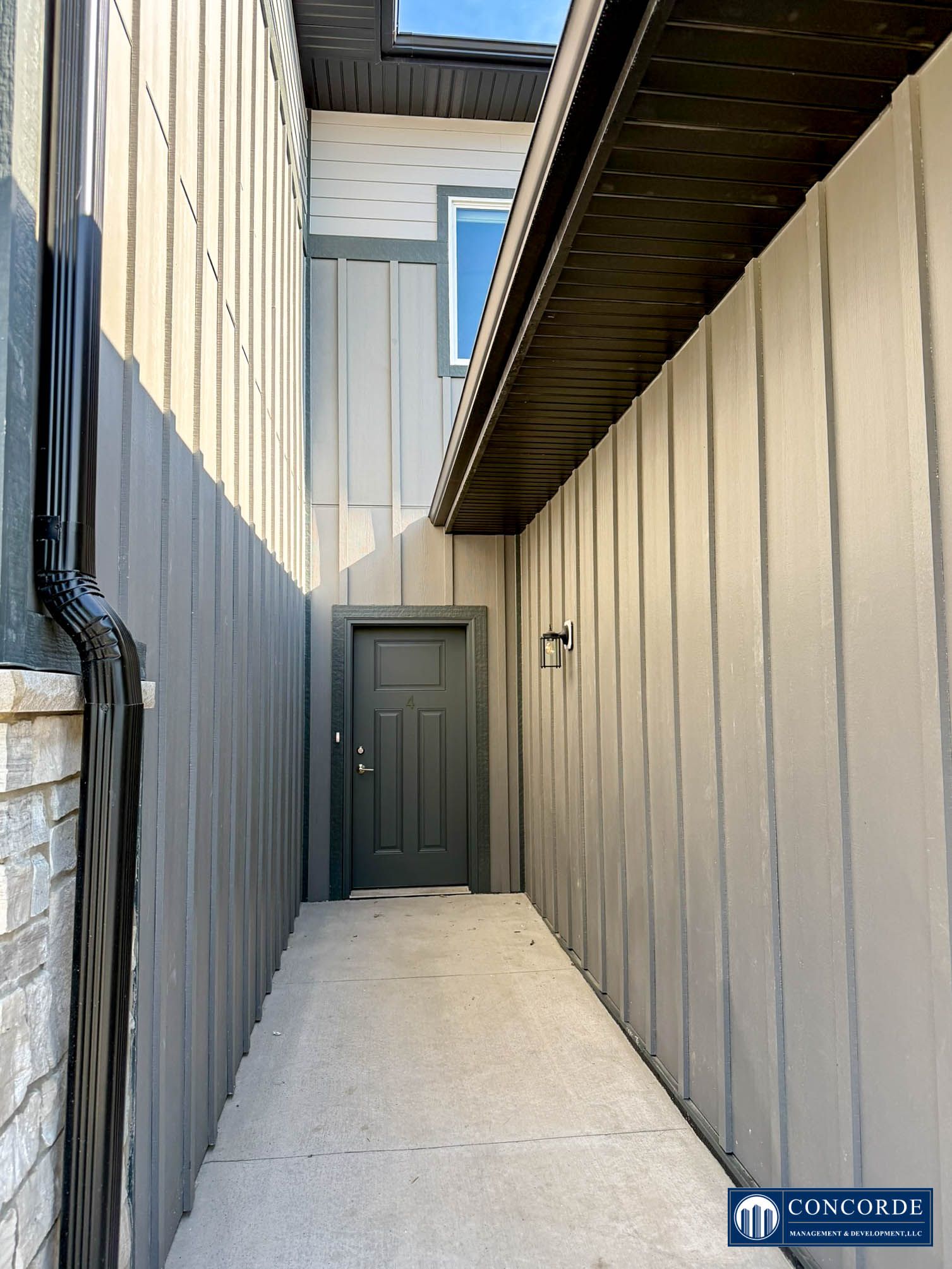 Narrow outdoor side passage with beige siding, white concrete path, and a gray door at the end