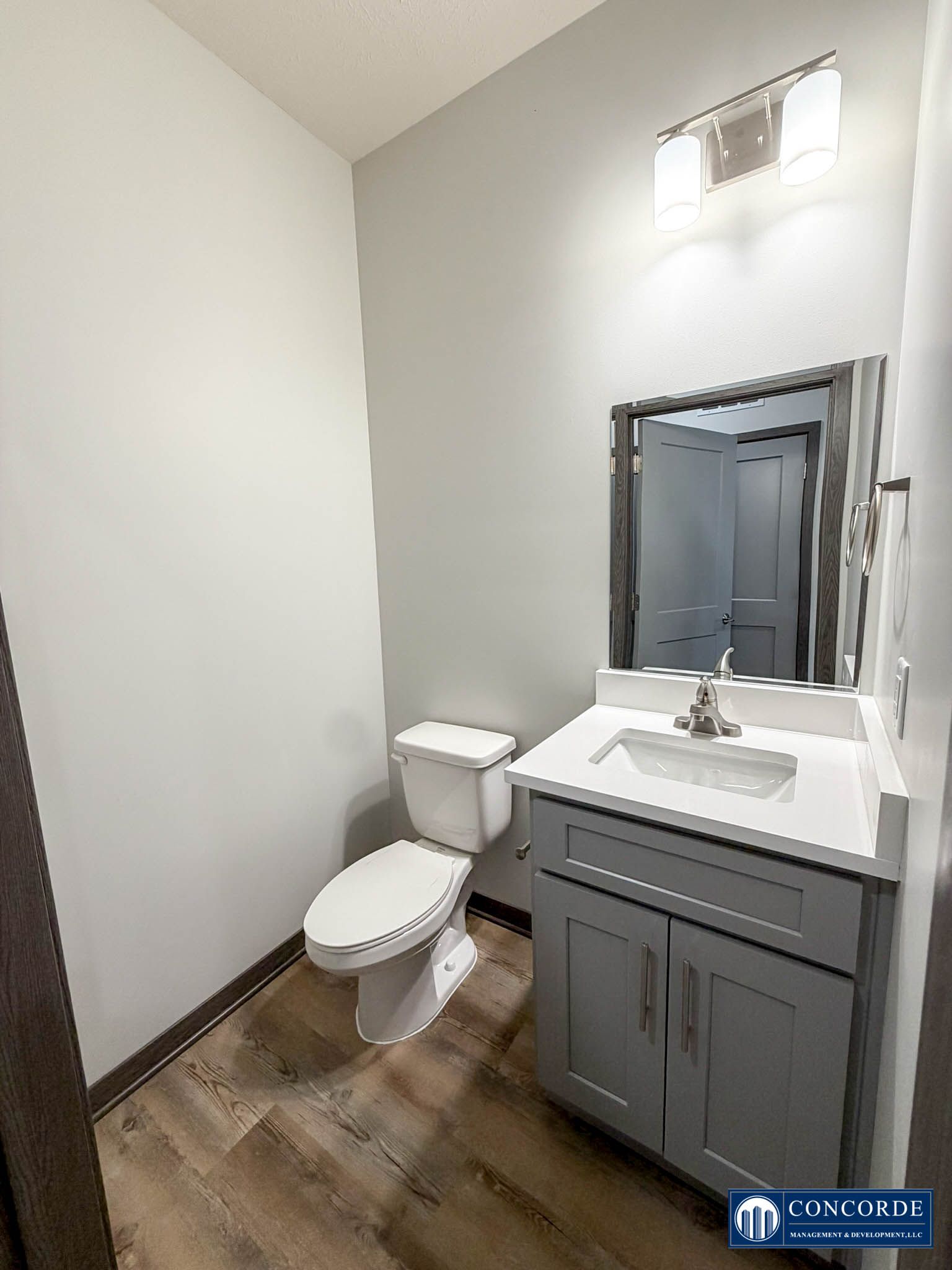 Small bathroom with toilet, vanity sink, mirror, wood-look floor, and frosted window.