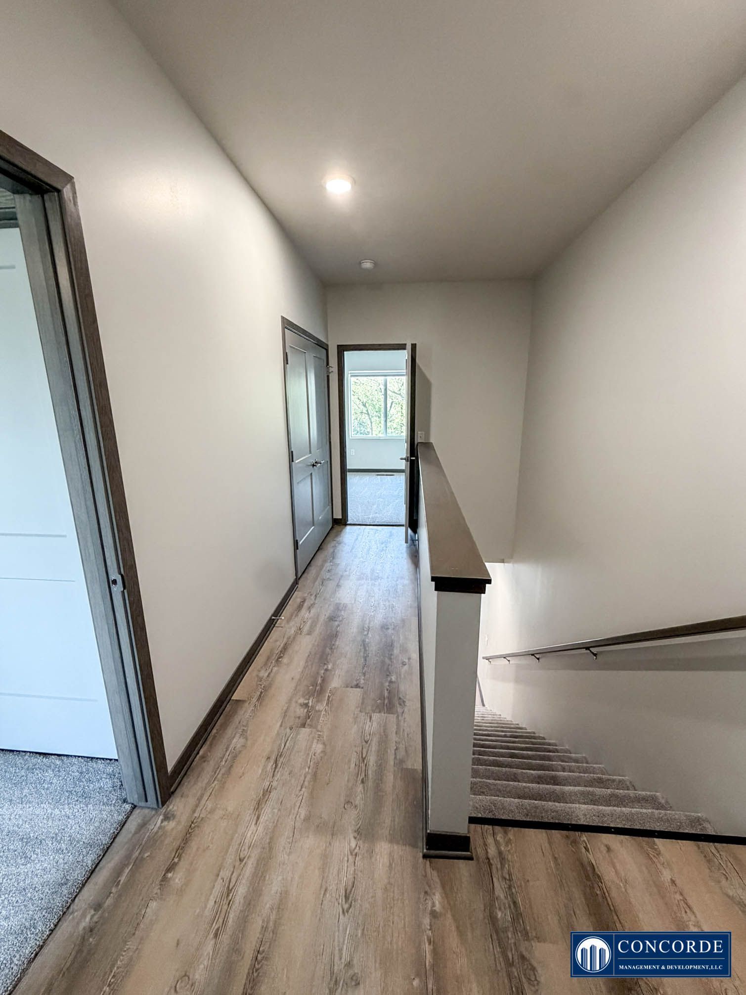 Hallway with wood flooring, white walls, stair railing, and a door opening to daylight.