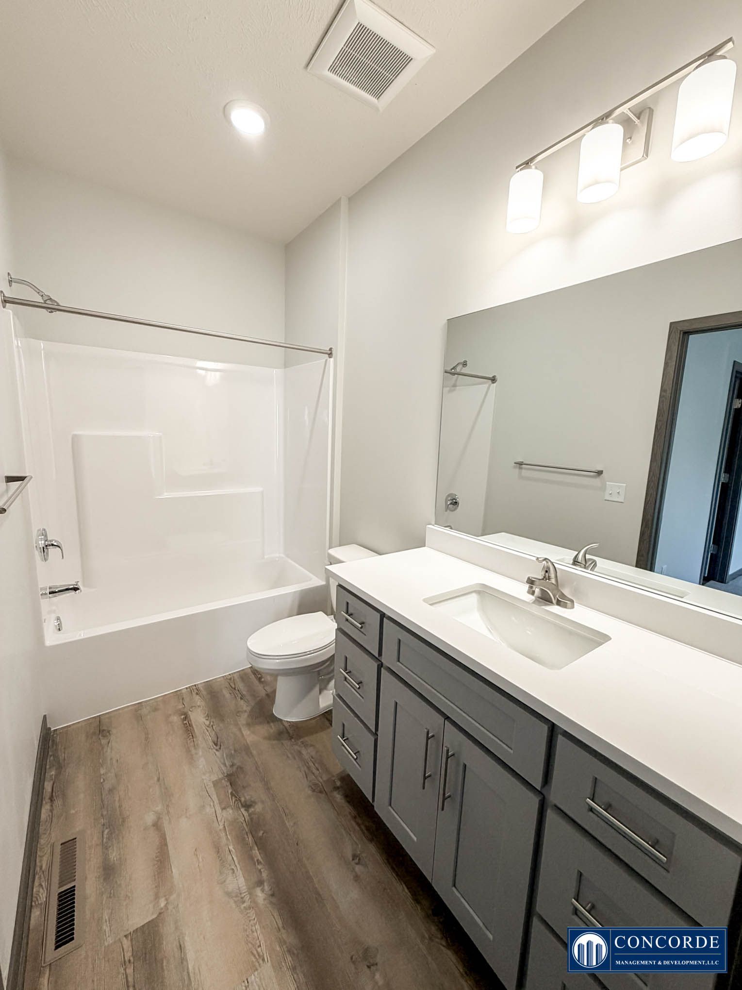 Modern bathroom with white tub, toilet, large mirror, and gray vanity on wood-look flooring